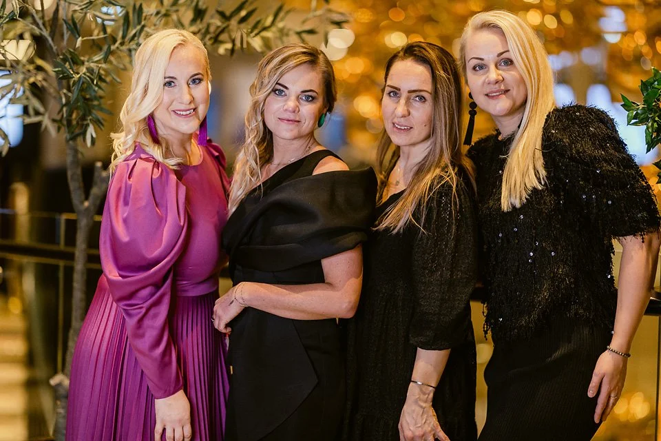 Four women in formal attire pose together at an indoor event with warm lighting and decorative plants in the background.