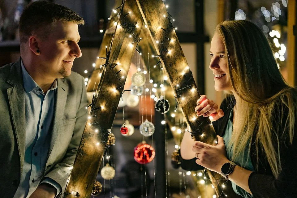 A man and a woman are looking at each other through a wooden star-shaped decoration wrapped with string lights and decorated with Christmas ornaments, during a festive holiday celebration.