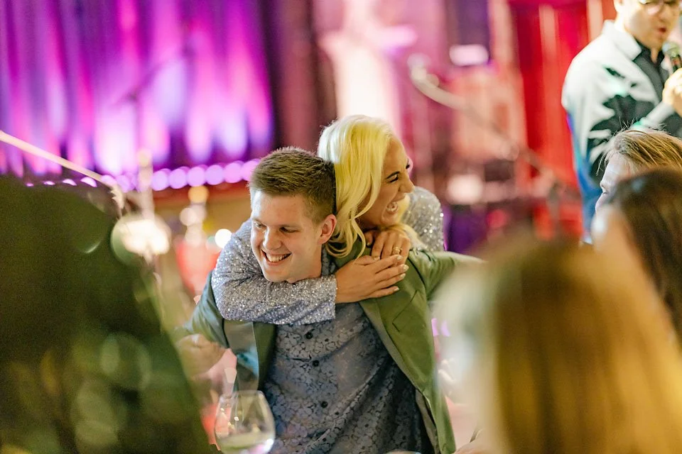 A man giving a woman a piggyback ride at a lively party, both smiling and enjoying the moment, with colorful lighting in the background.