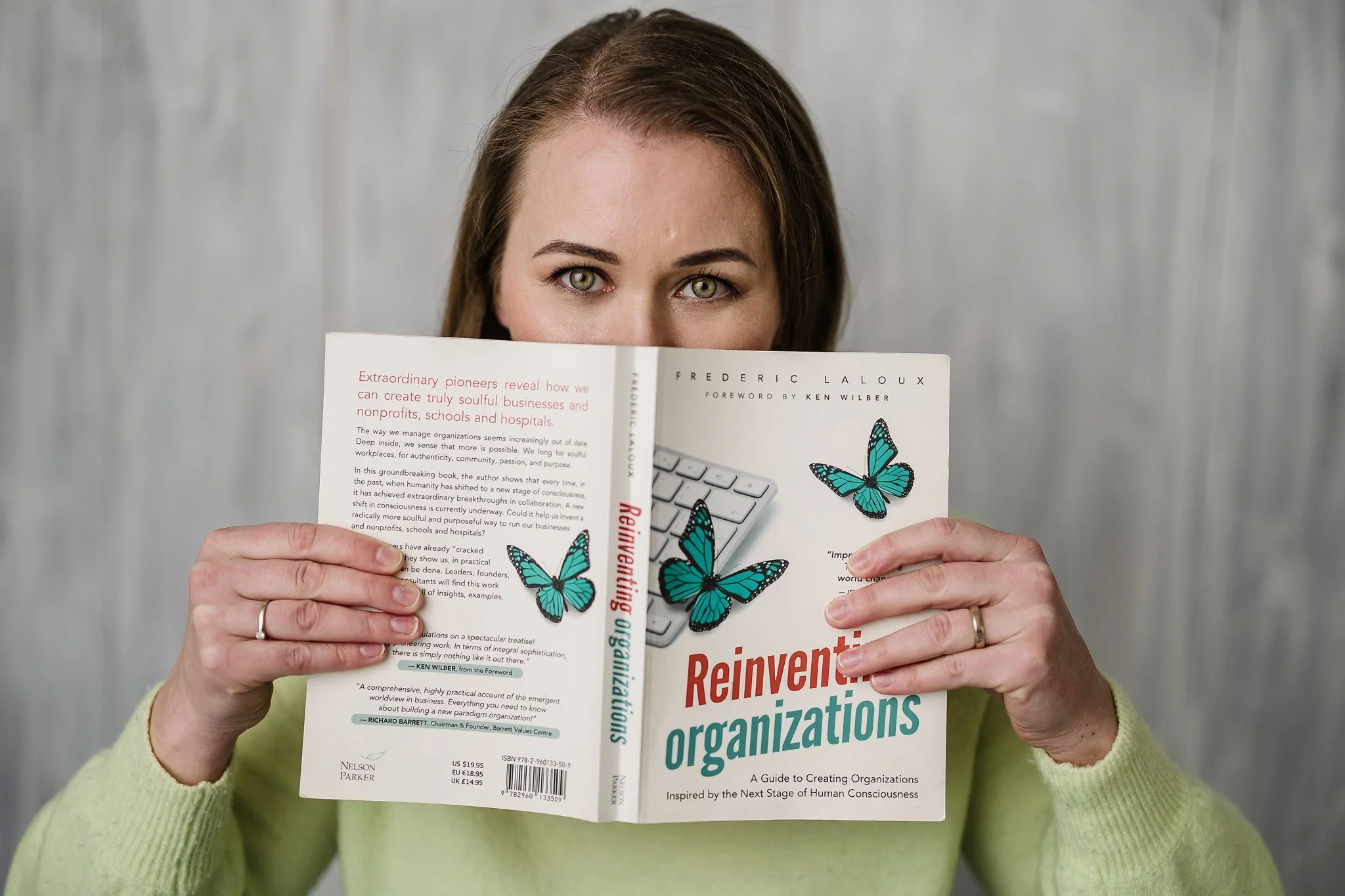 A woman with green eyes and light brown hair holding a book titled "Reinvent Organizations" in front of her face, revealing only her eyes, with a gray background.