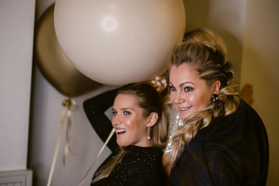 Two women smiling at a celebration, decorated with large gold and white balloons in the background.