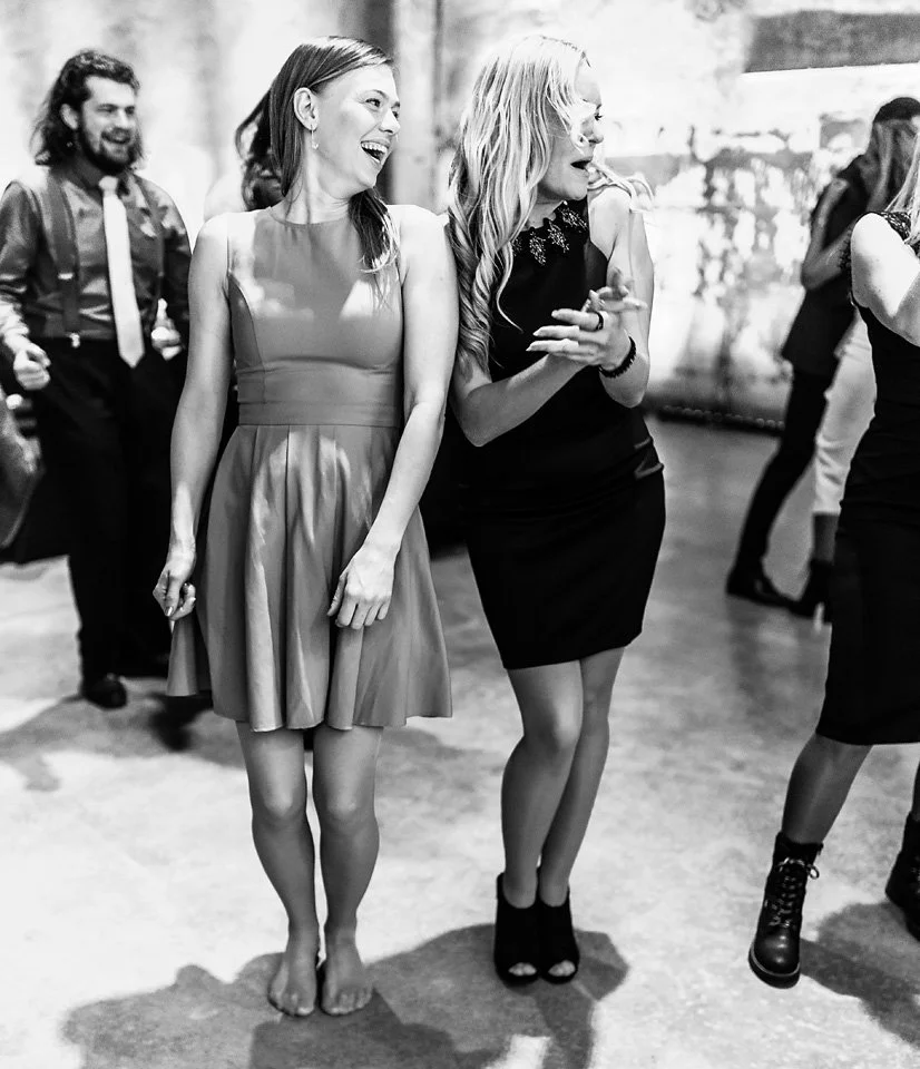 Two women dressed in elegant attire laughing and enjoying themselves at a social gathering, with other guests in the background.