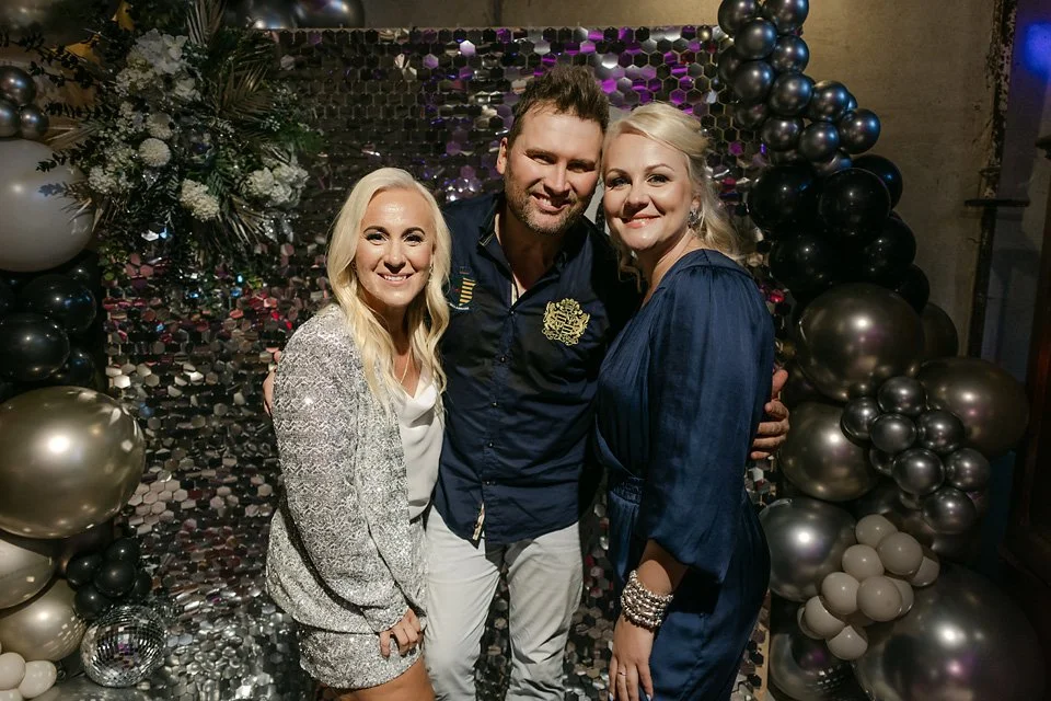 Three people, two women and a man, smiling and posing together at a celebration with balloon decorations and a floral backdrop.