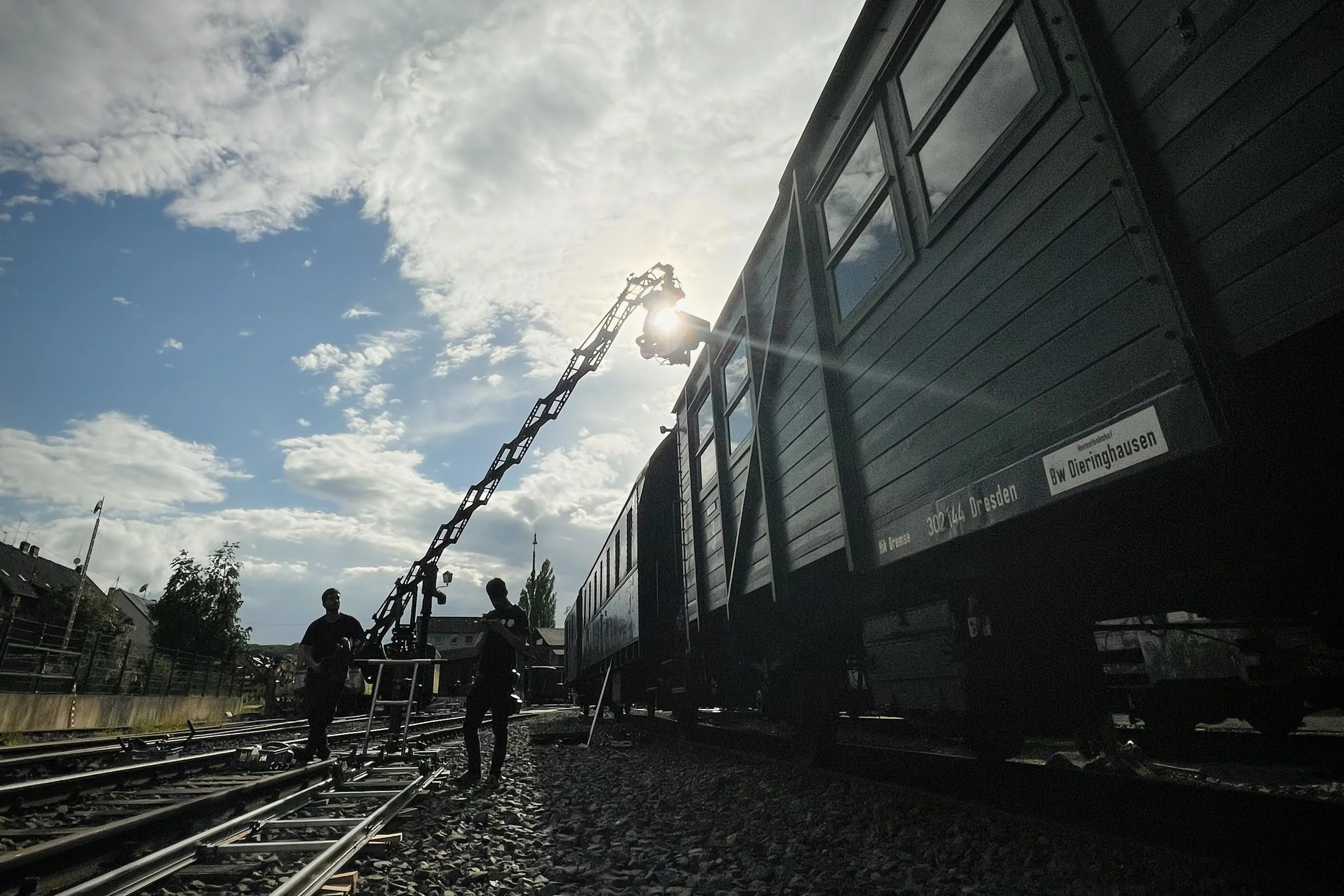 Two workers working on railway tracks near a train car, with a crane lifting a part, under a partly cloudy sky during daytime.