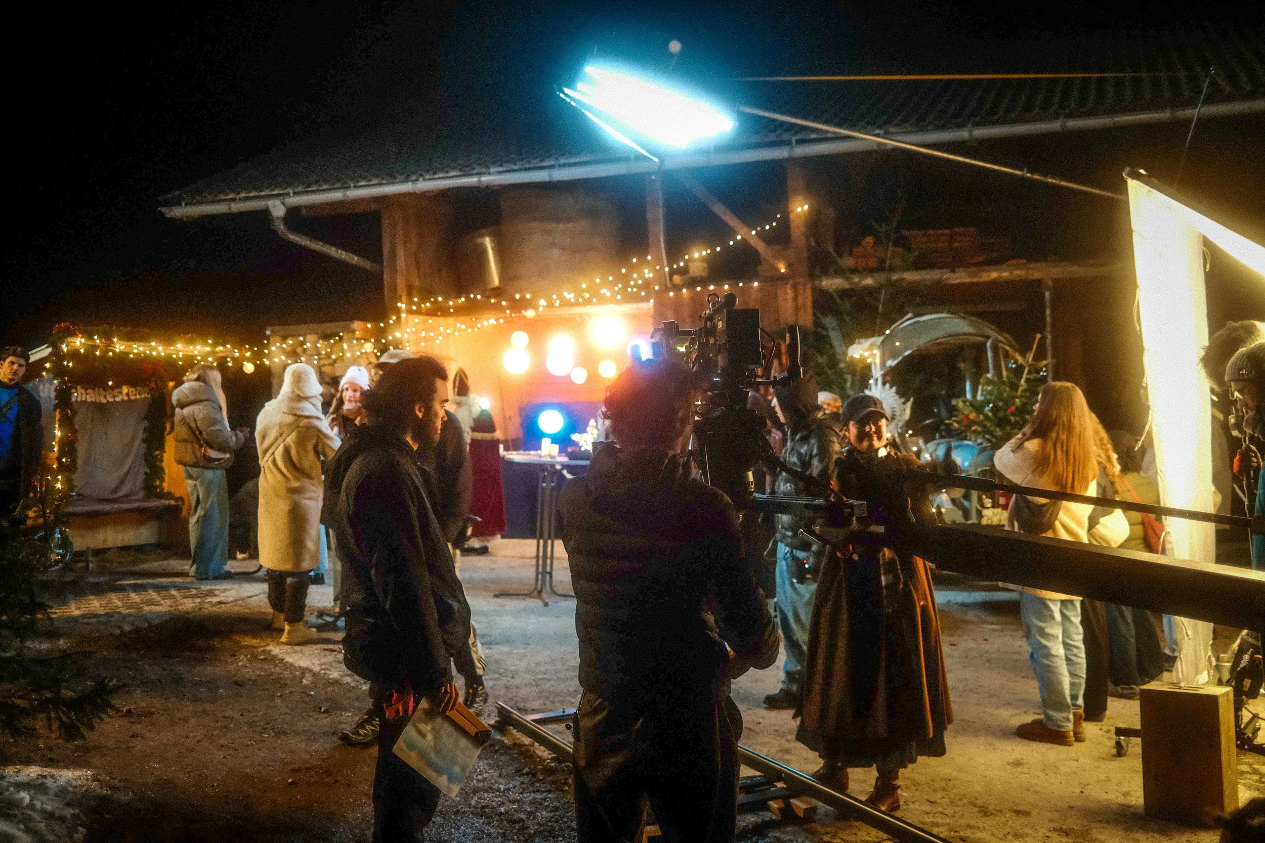 People gathered outside a rustic building decorated with Christmas lights at night, with a camera crew filming a scene or event.