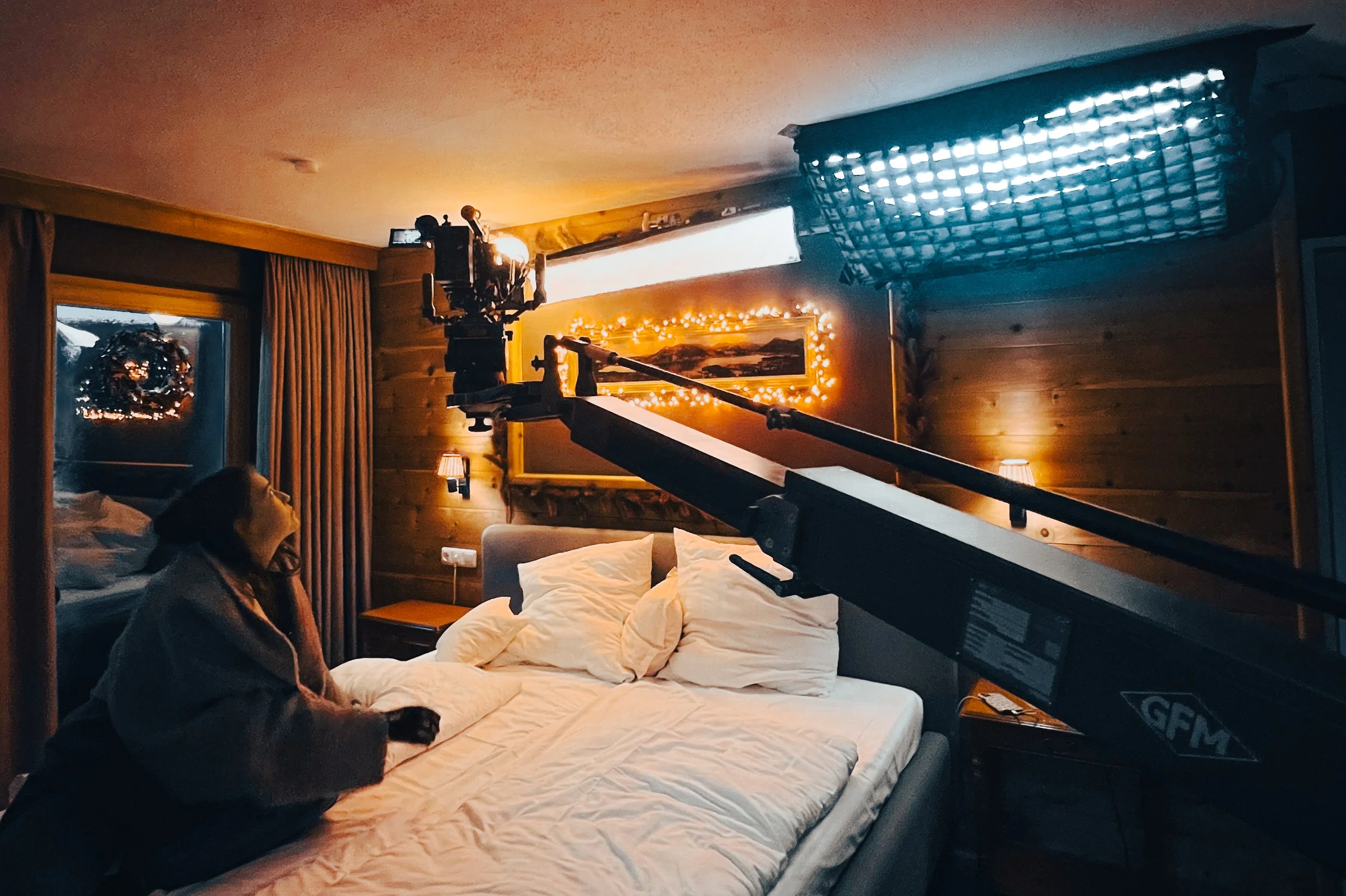A person sitting on a bed in a wooden-paneled bedroom with Christmas lights, a wreath in the window, and filming equipment pointed at them.