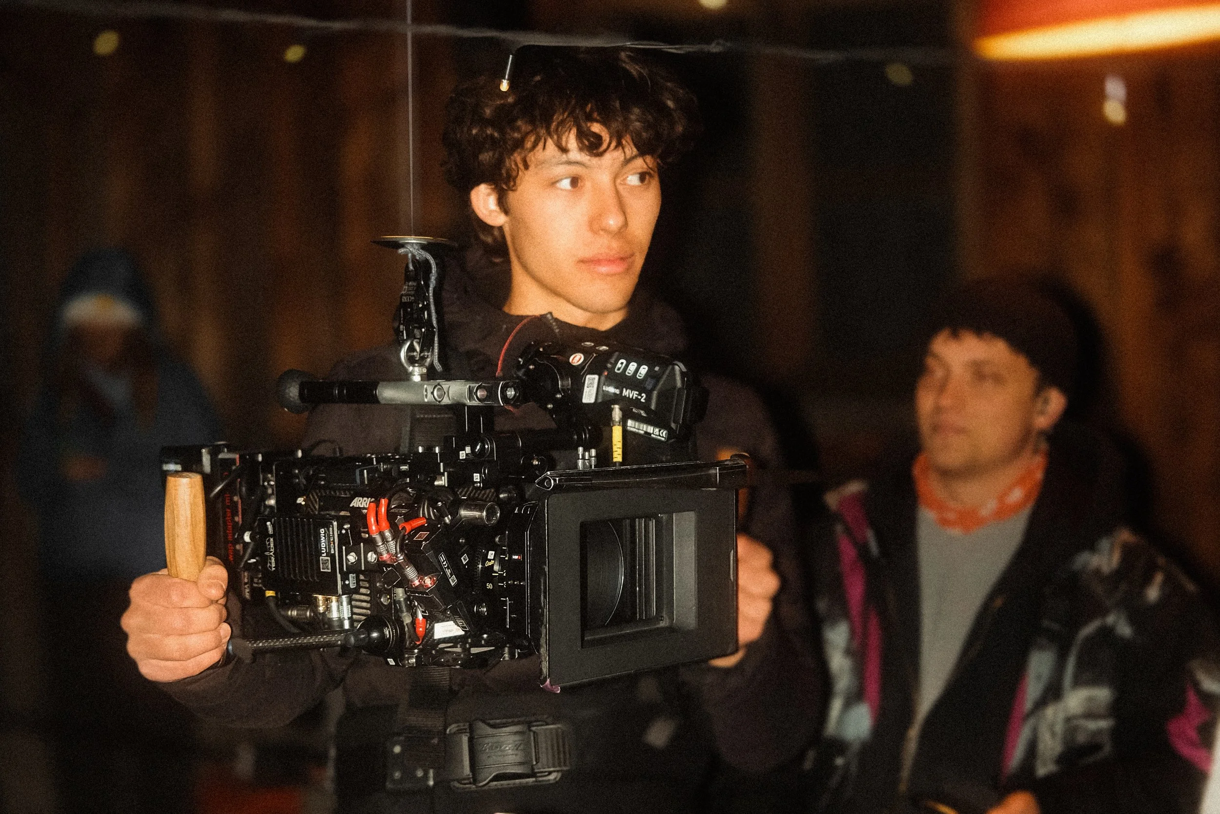 Young man holding professional film camera on set, with others in background.
