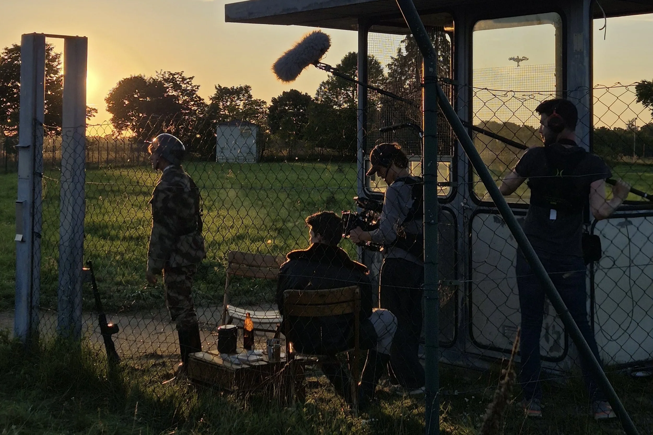 A film crew is filming a scene outdoors during sunset, with one person in costume inside a fenced area and others operating equipment outside the fence.