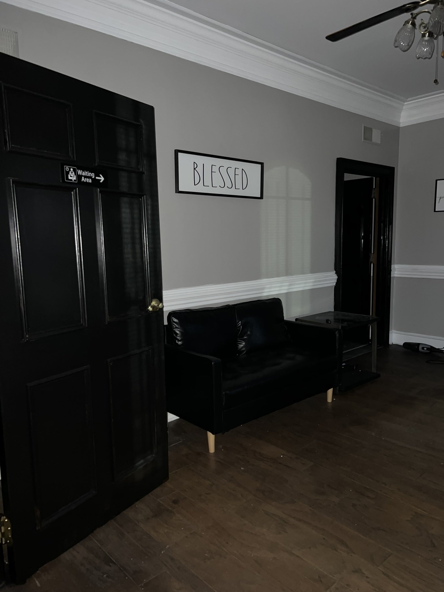 Interior of a room with gray walls and white trim, featuring a black door labeled 'Waiting Area' with a gold handle, a black sofa with wooden legs, a black table, and framed wall decor with the word 'BLESSED.'
