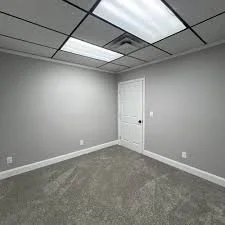 Empty room with gray walls, white door, ceiling tiles, and beige carpet flooring.