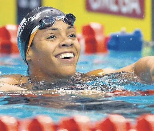 A female swimmer in a swimming pool, wearing a swim cap and goggles, smiling after completing her race.