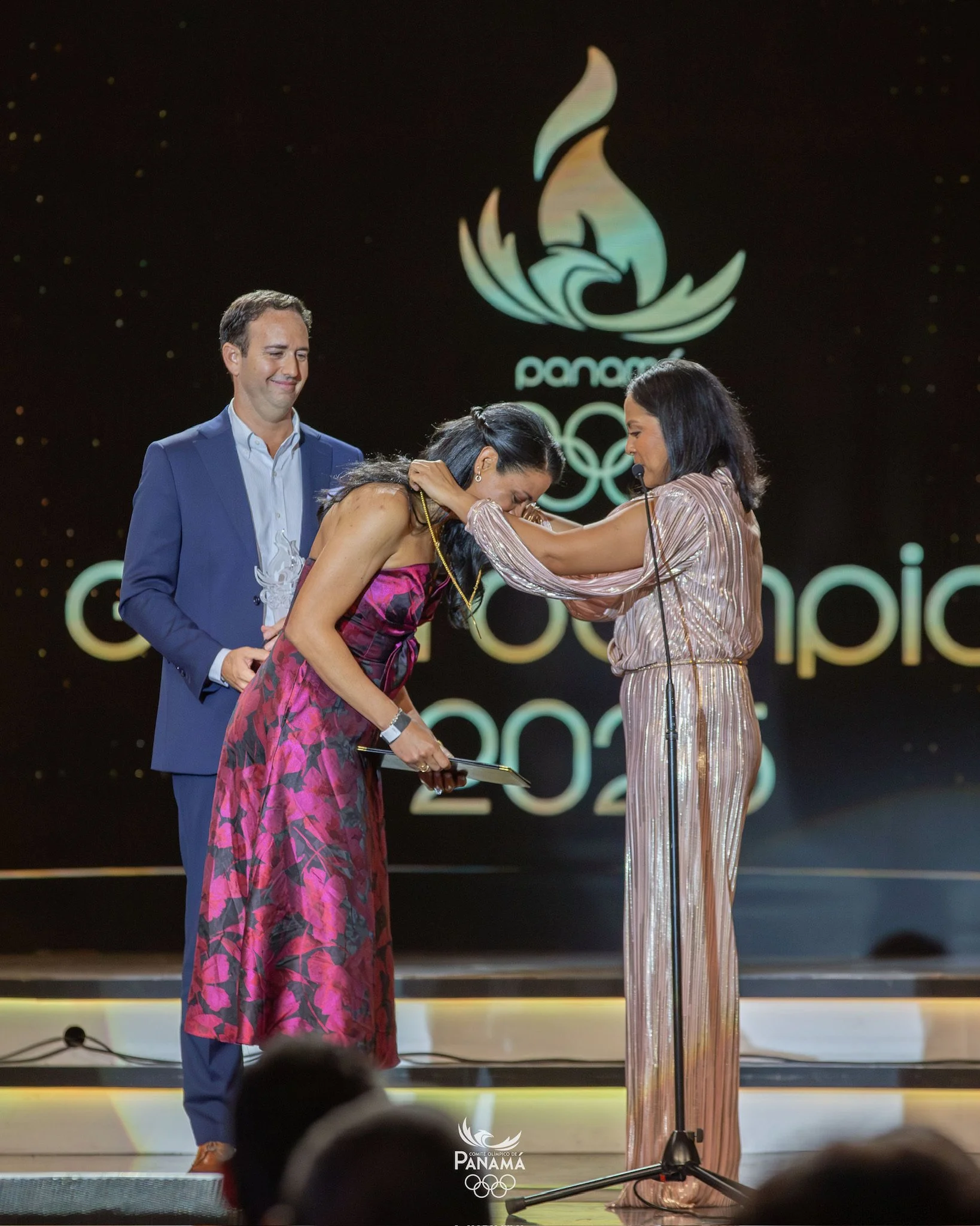 An award ceremony on stage with three people: a woman in a pink floral dress receiving a medal, a woman in a shiny striped outfit presenting the medal, and a man in a blue suit standing behind them. The background shows a logo and the words 'Panama 2023'.