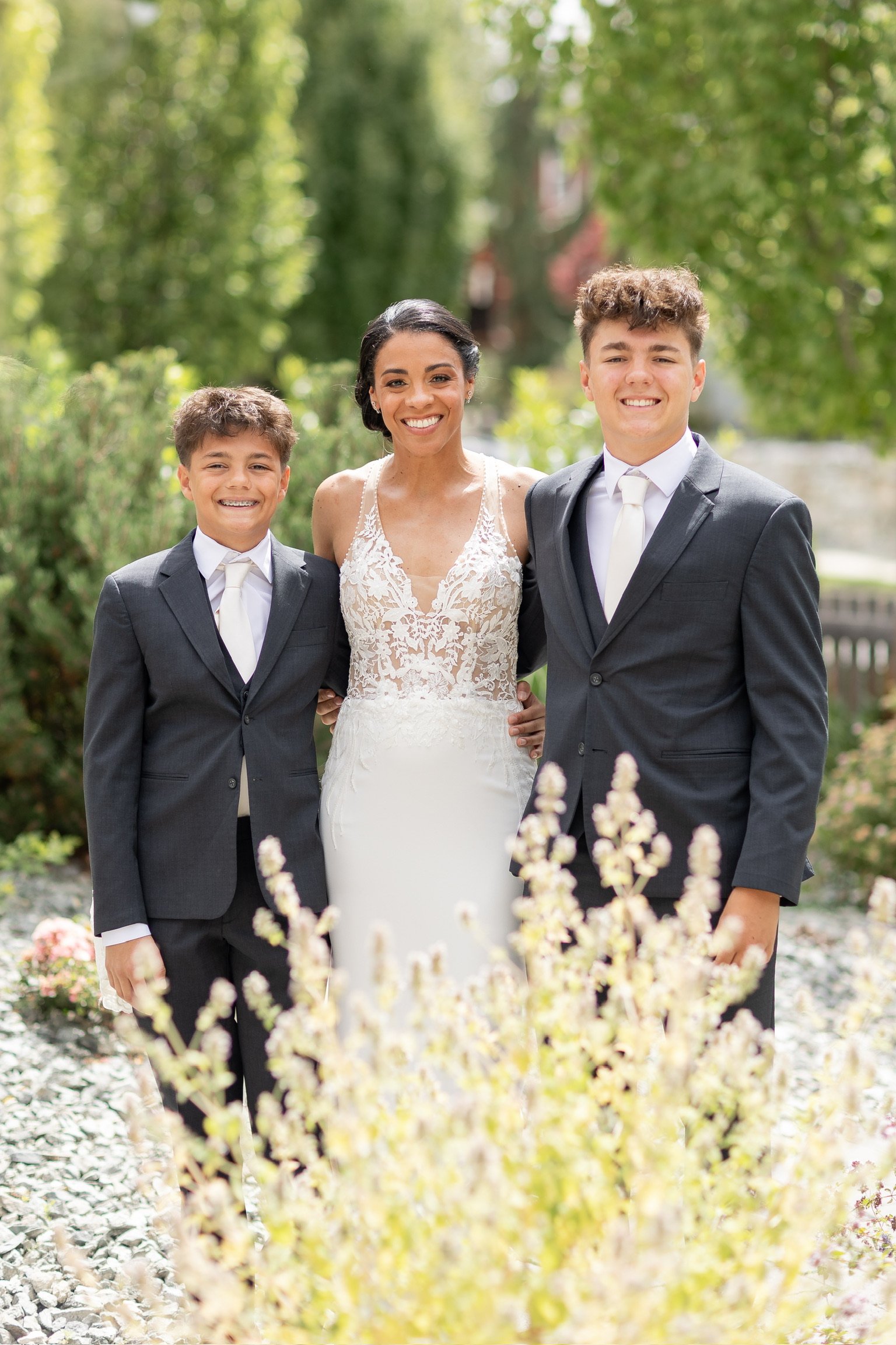 A woman in a white lace wedding dress standing between two boys in suits outdoors. All smiling in a garden with trees and flowers.