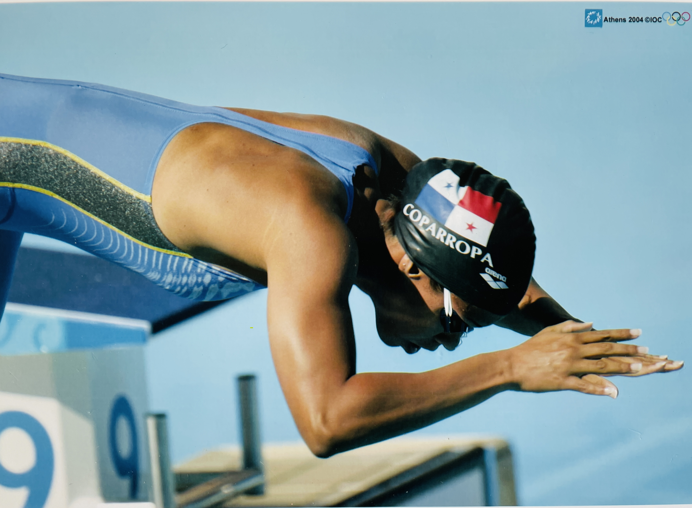 An athlete in a blue swimming suit and black swimming cap crouches on a starting block, preparing to dive into the pool during a race at the Athens 2004 Olympics.