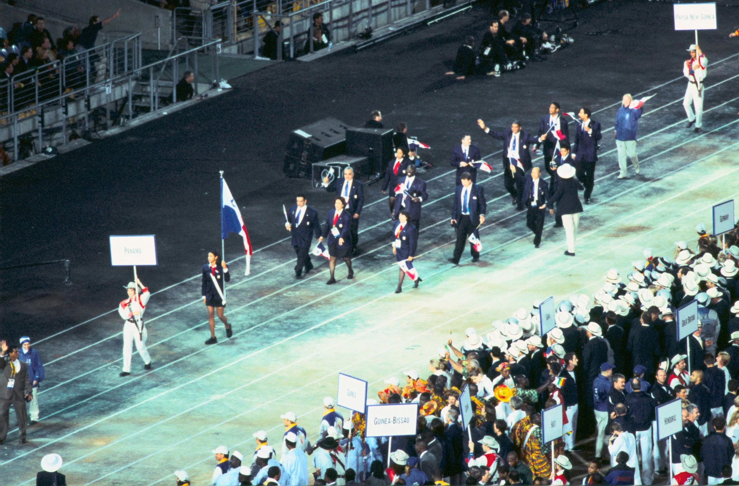 People marching in a parade with flags and signs representing different countries, while a seated audience watches.