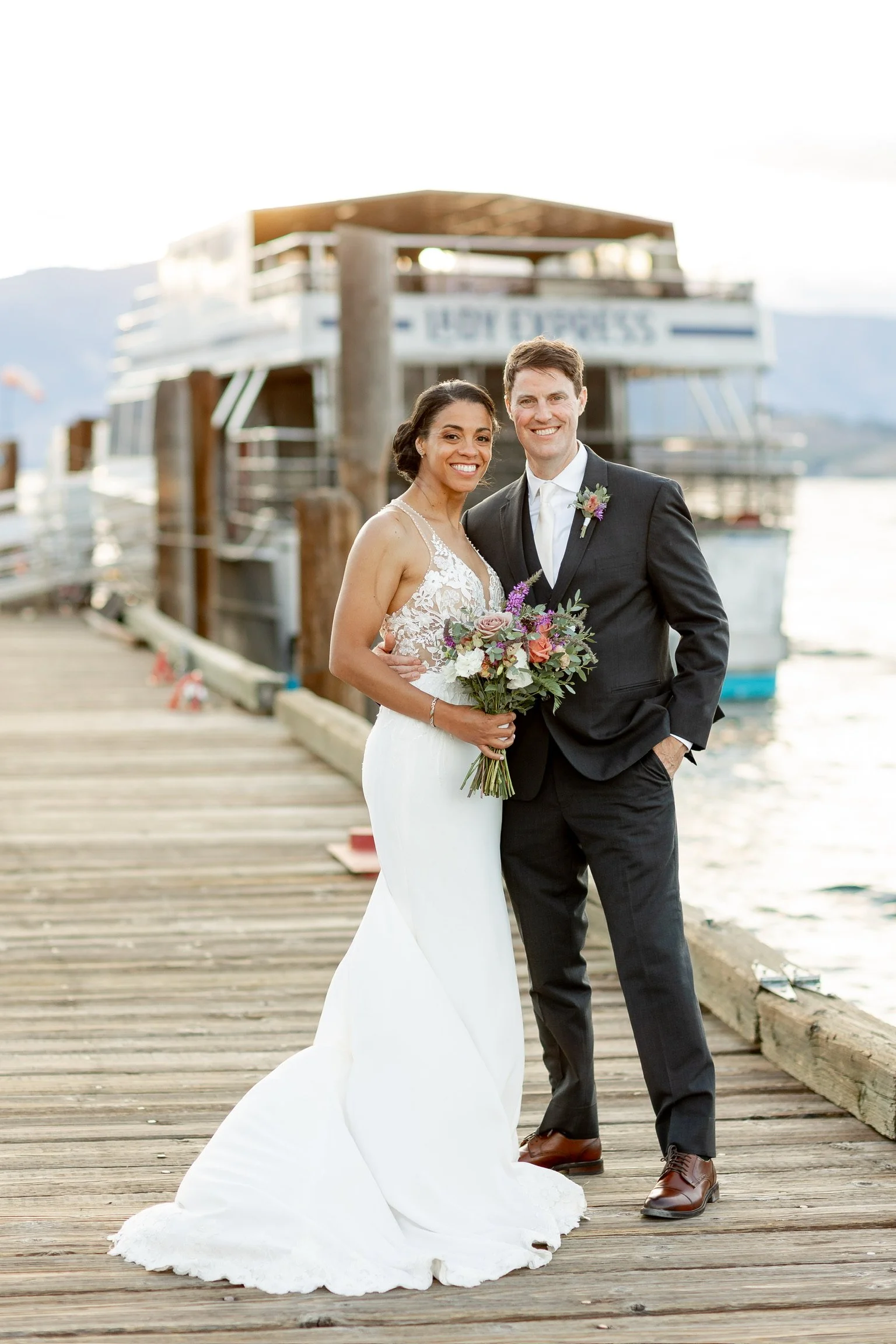 A newlywed couple standing on a wooden dock by the water, smiling, with a boat docked behind them. The bride is holding a bouquet of flowers, and they are dressed in wedding attire.