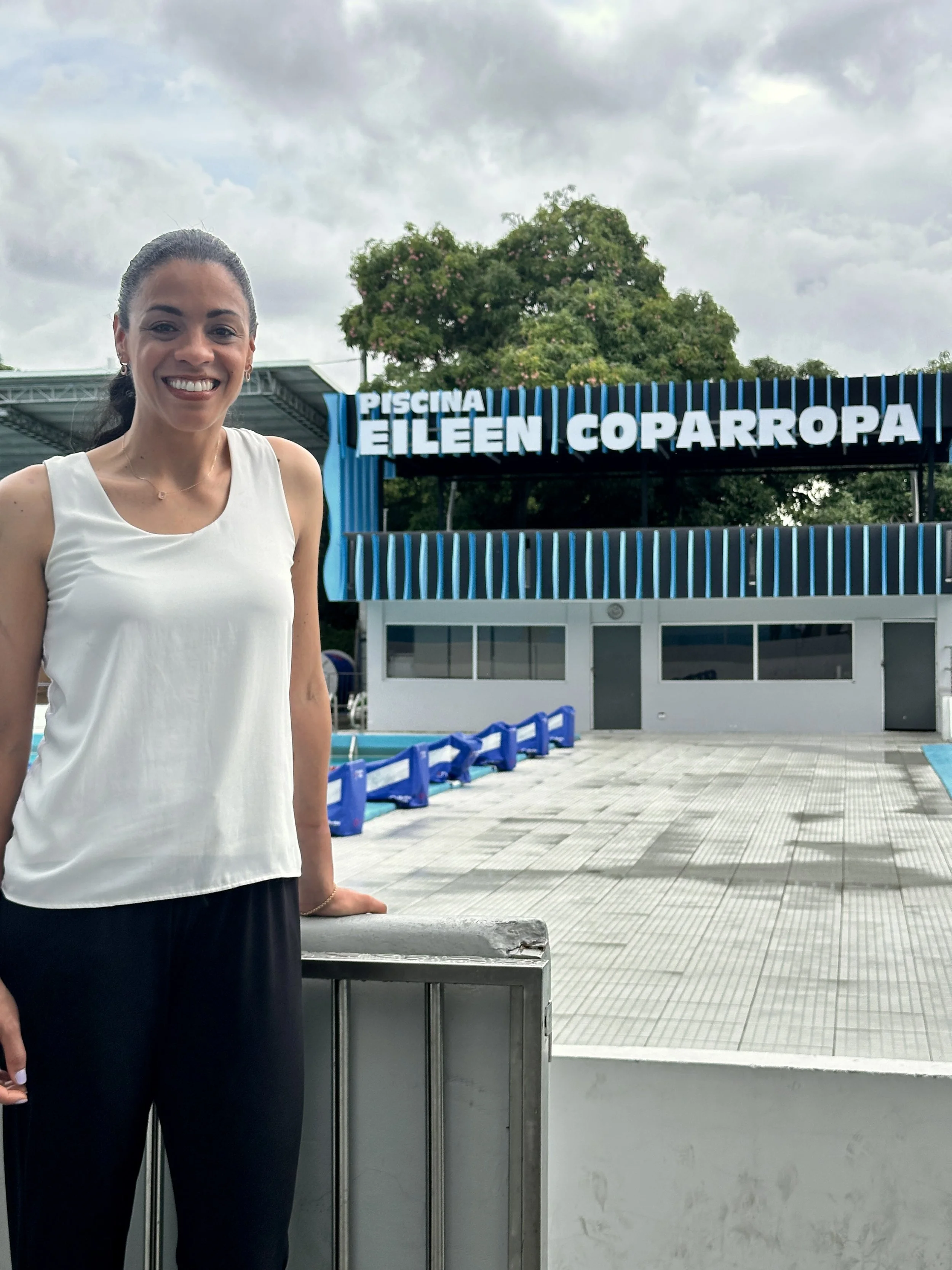 A woman in a white sleeveless top and black pants standing outside a swimming pool and clubhouse at Piscina Eileen Coparropa, with cloudy sky and trees in the background.