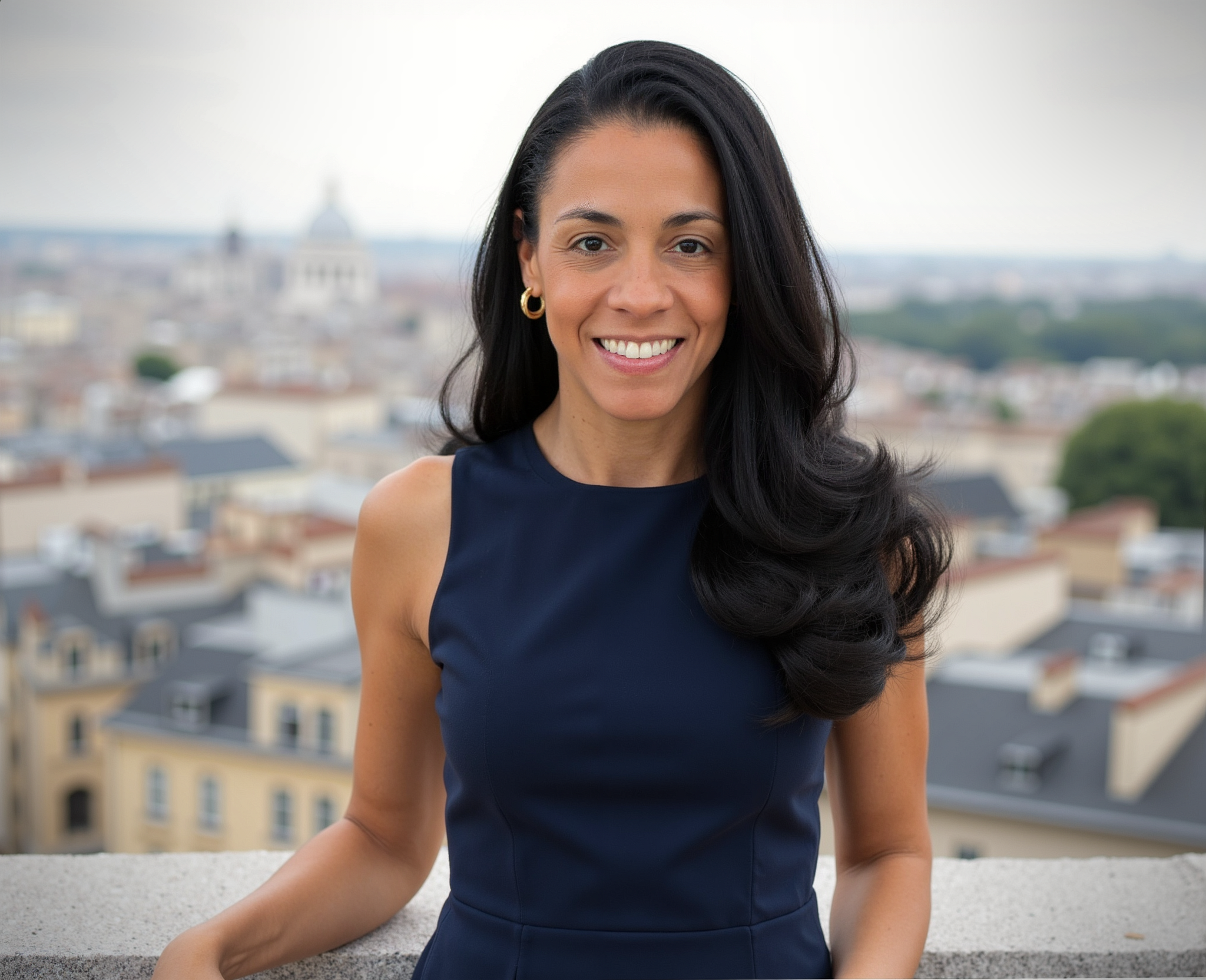 Woman with long dark hair smiling, wearing a navy sleeveless dress, standing outdoors with city rooftops and a domed building in the background.