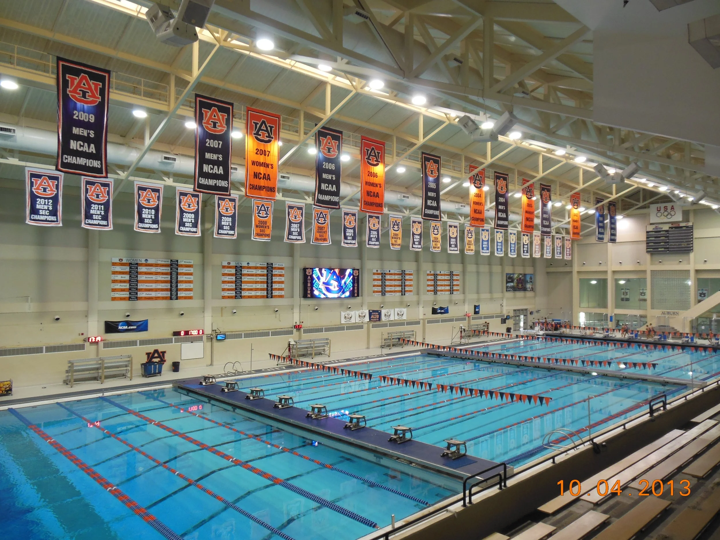 Indoor swimming pool with multiple lanes, banners of Auburn University celebrating sports championships, and large digital scoreboard, dated October 4, 2013.