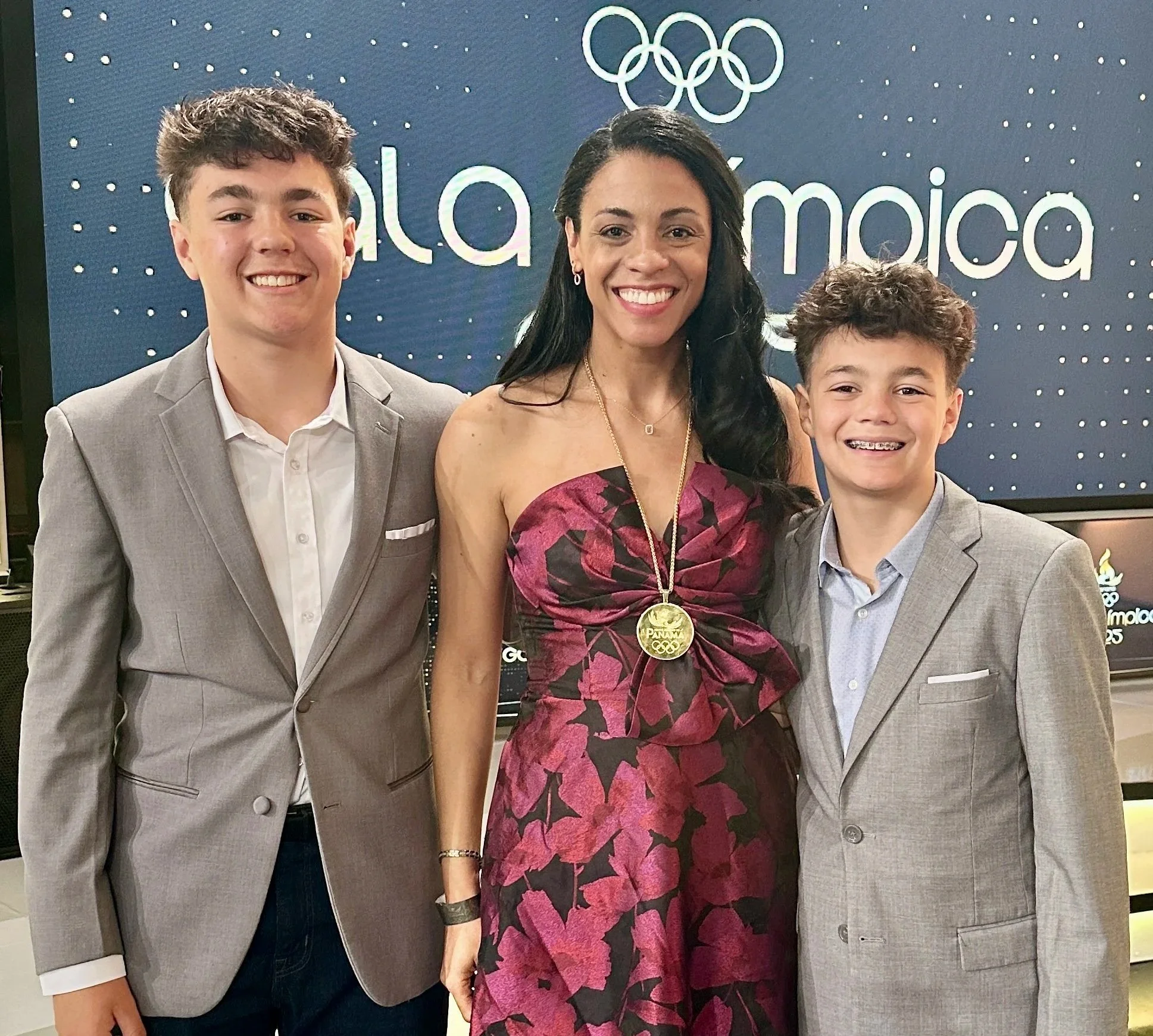 A woman and two young boys in suits at the Tokyo 2020 Olympics, smiling in front of a blue background with the Olympic rings and the word Palma.