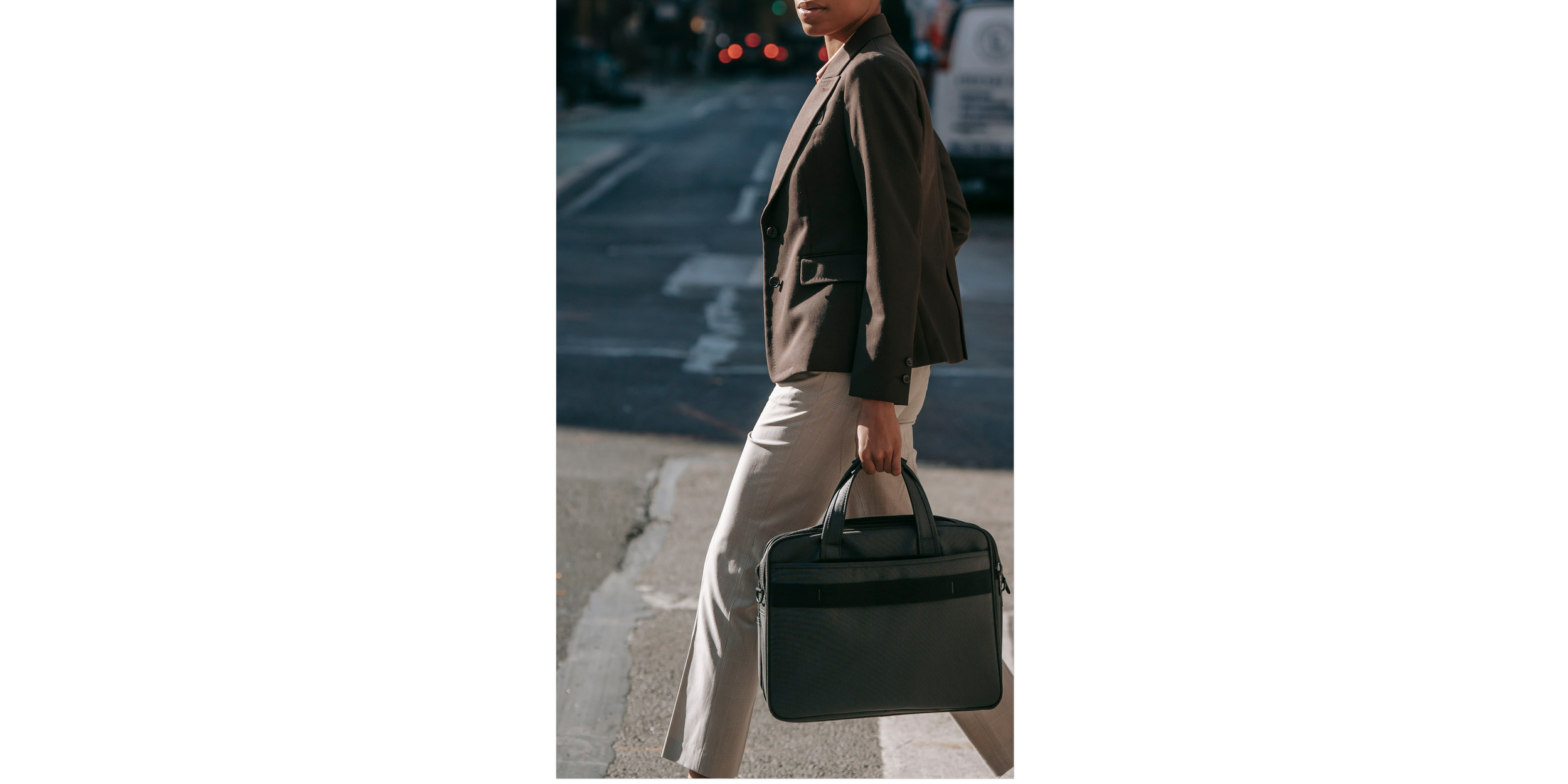A person wearing a brown blazer and beige pants standing on the street, holding a black briefcase or laptop bag in their right hand.