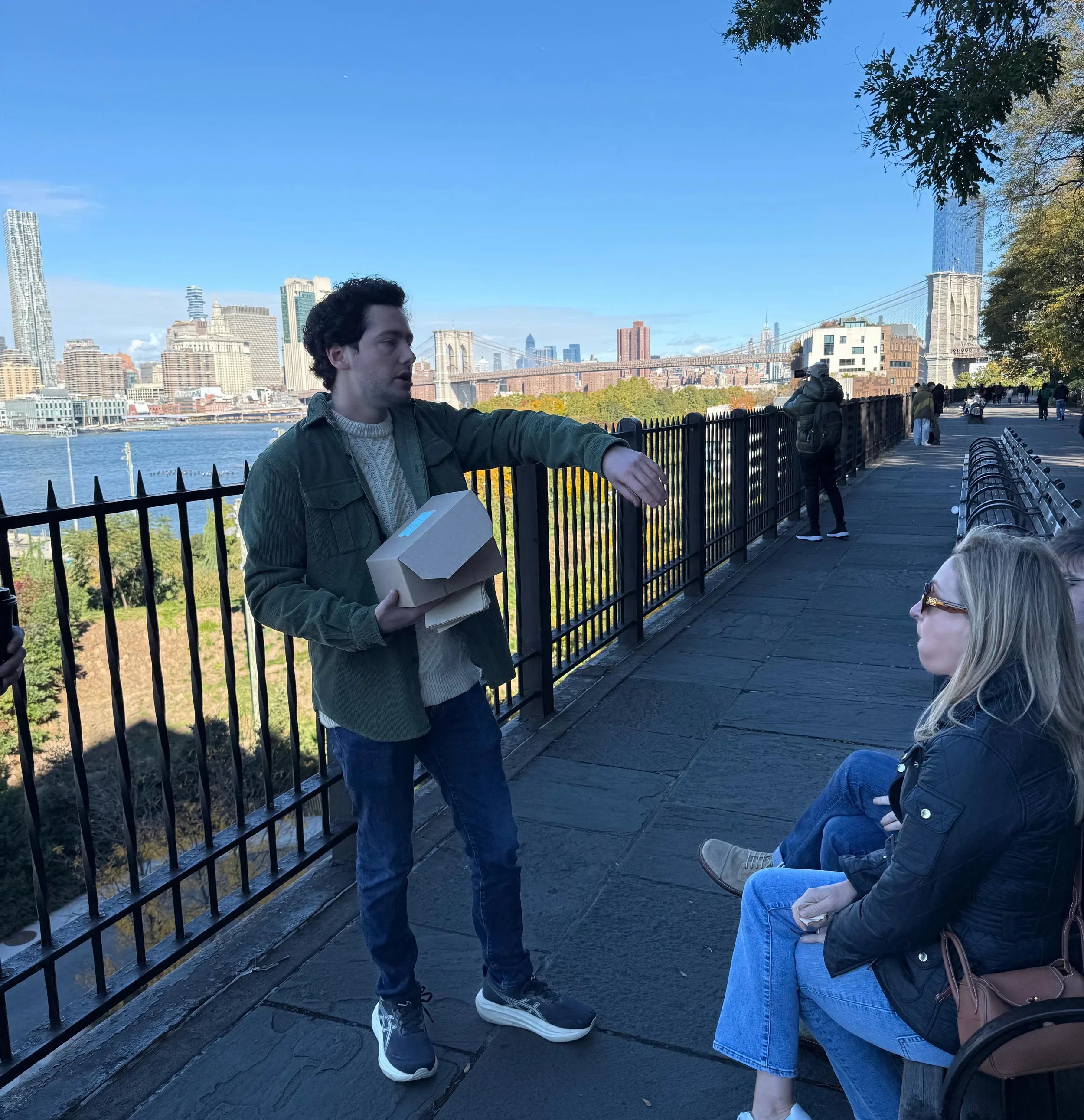 A young man is talking to two women sitting on a park bench near the waterfront with a city skyline and bridge in the background.