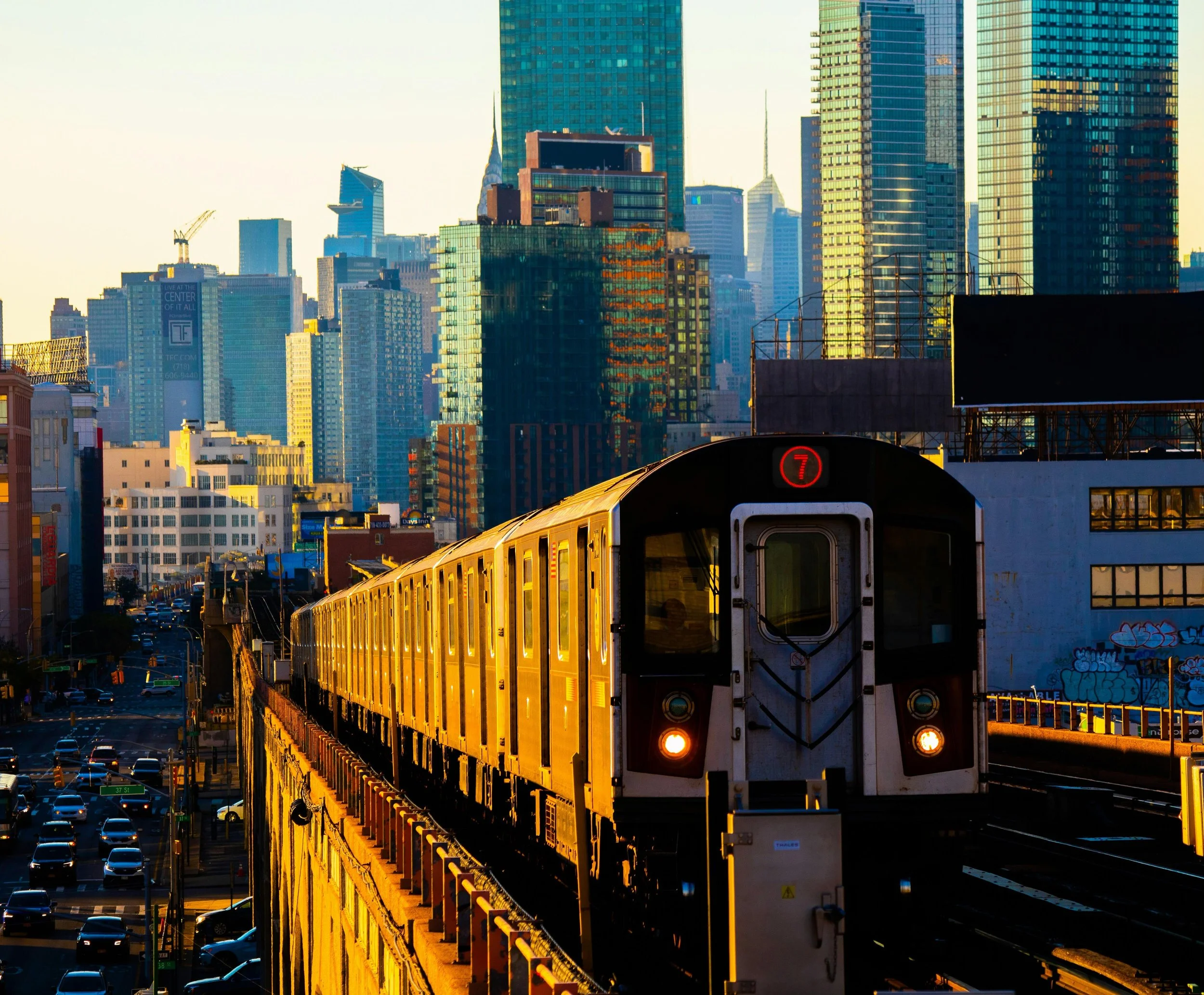 A yellow train traveling on an elevated track through a city with tall skyscrapers in the background during sunset.