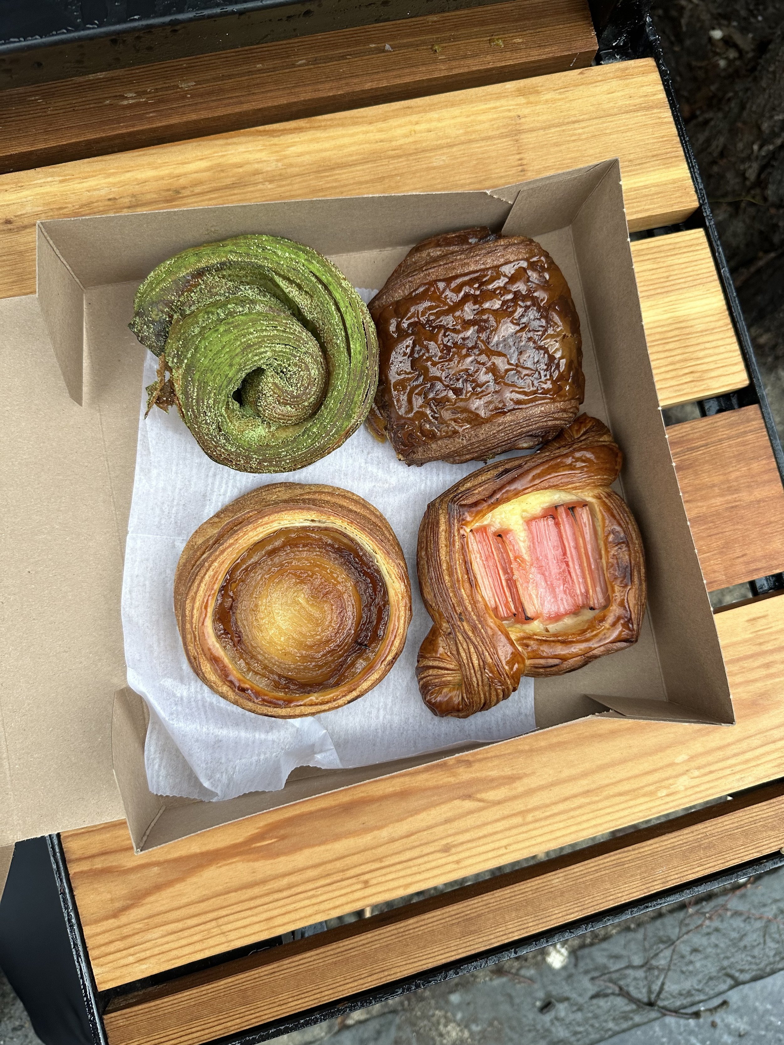 A box of four assorted pastries on a wooden surface, including a green spiral pastry, a square sticky bun, a caramel-topped pastry, and a pink-crusted Danish with a layered filling.