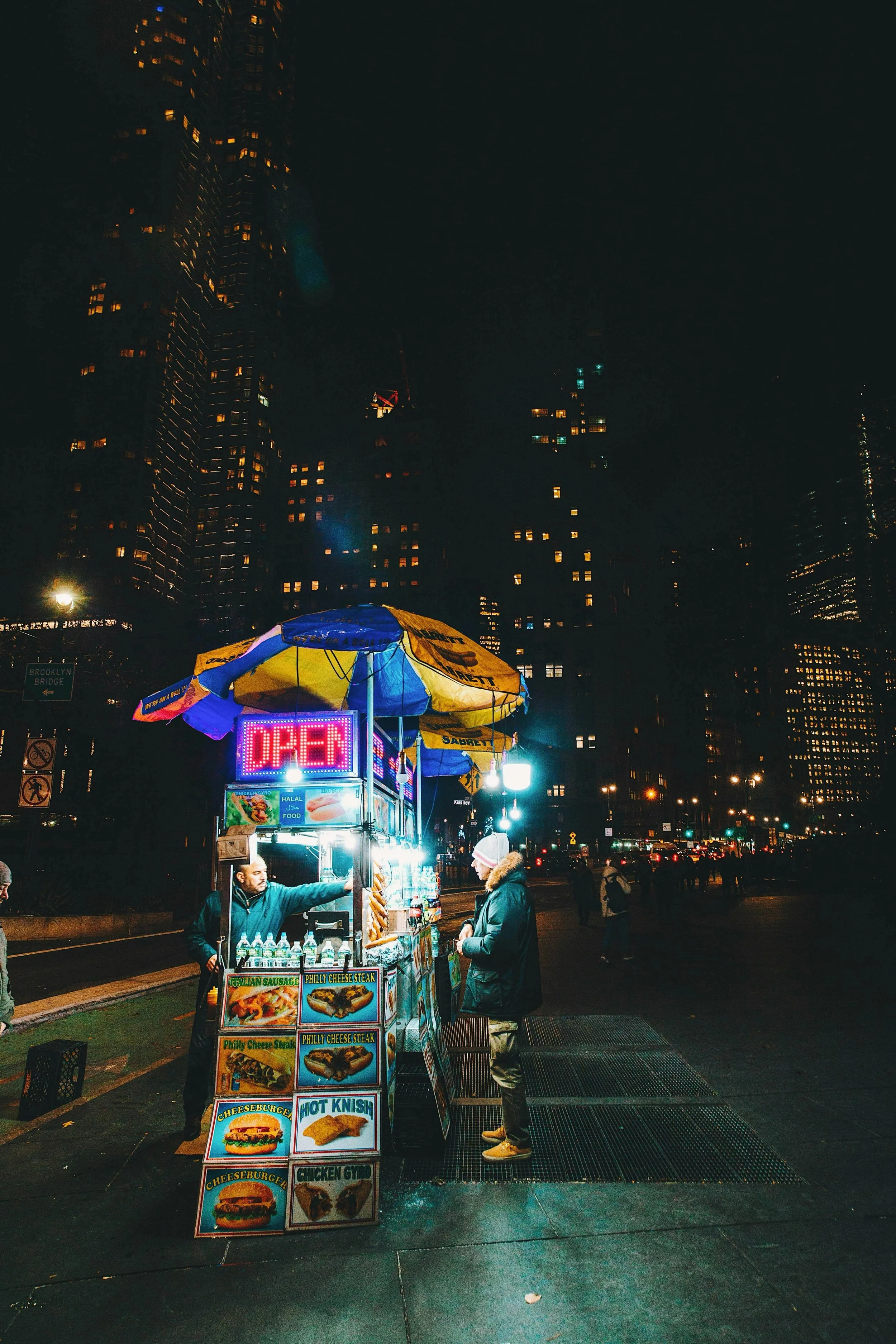 Night scene of a street food stand with a neon 'OPEN' sign, serving food to a customer, in front of tall city buildings with illuminated windows.