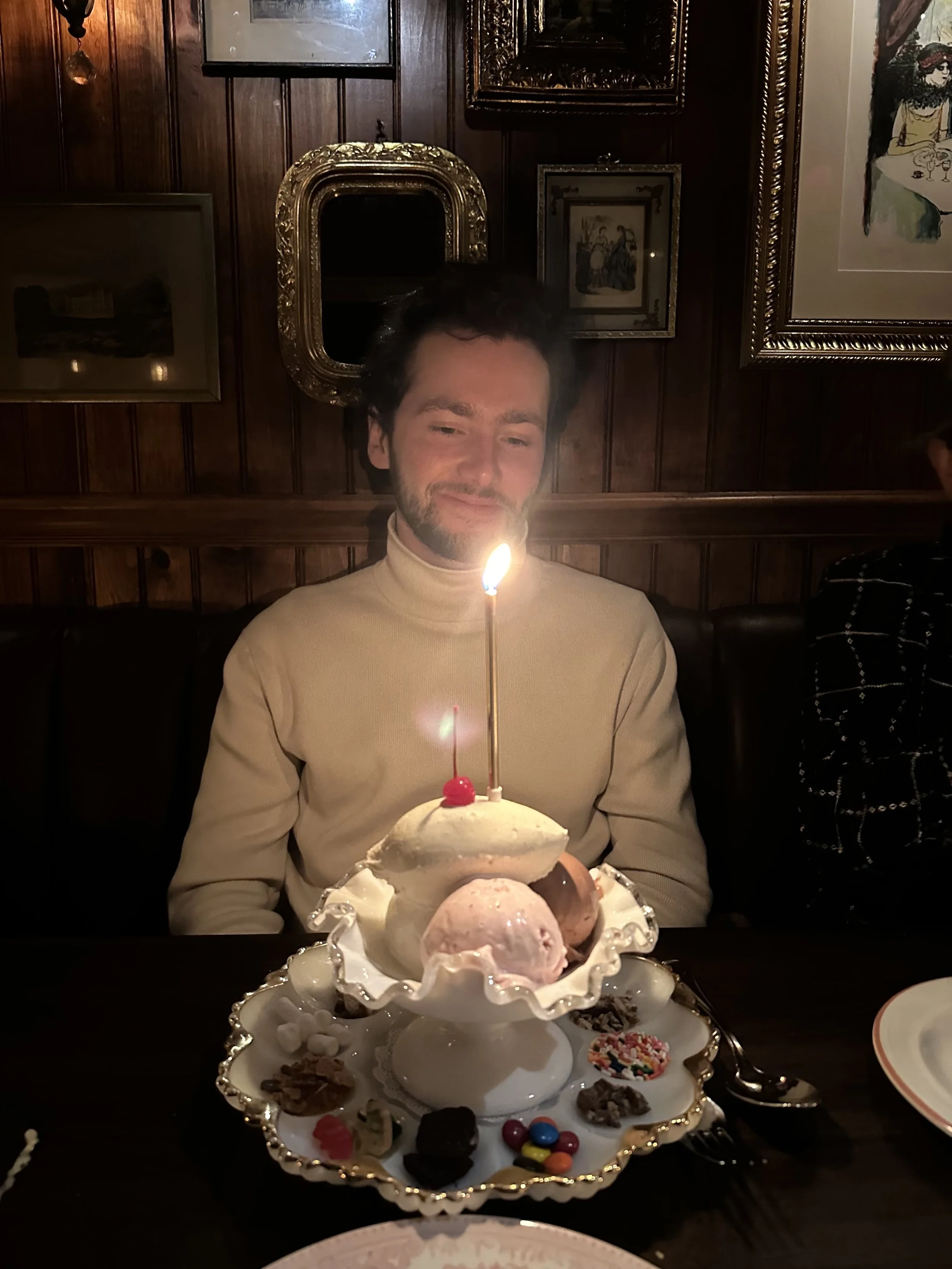 Man with dark hair and beard wearing a beige turtleneck sweater, sitting at a dark wooden table in a dimly lit room, smiling at a dessert with a lit candle on top.