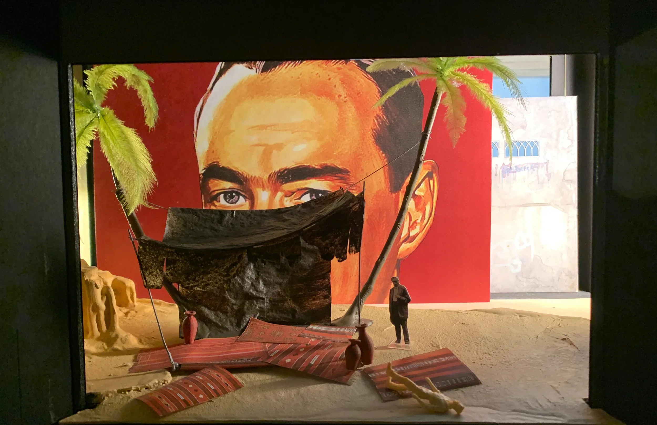 A giant face painted on the back wall peers over a makeshift tent strung between palm trees. Red rugs are strewn on the sand floor. One figure stands in the background, looking at another figure lying in the foreground.
