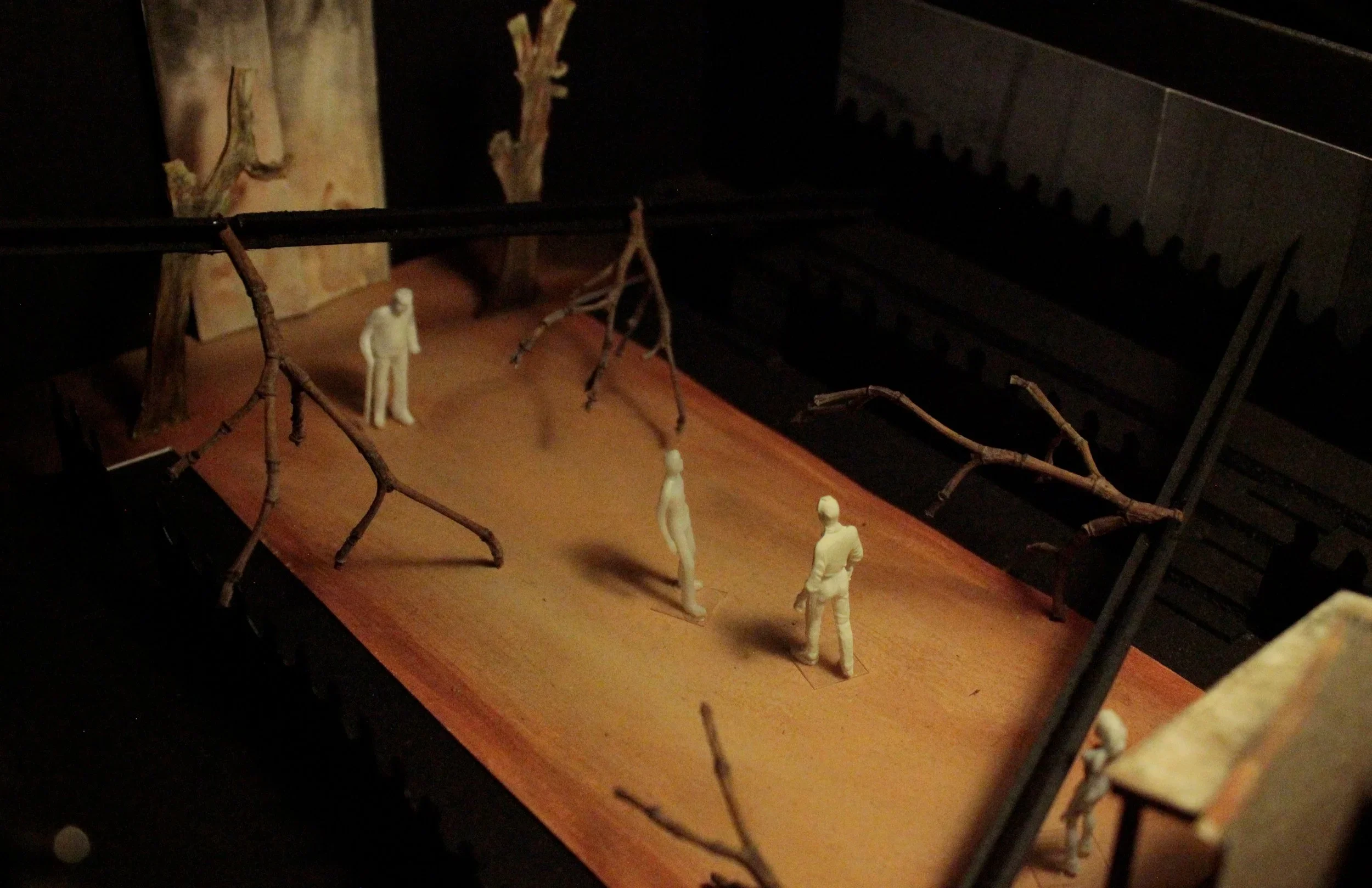 White plastic figures, surrounded by fallen tree branches, traverse a rectangular, packed-dirt floor.