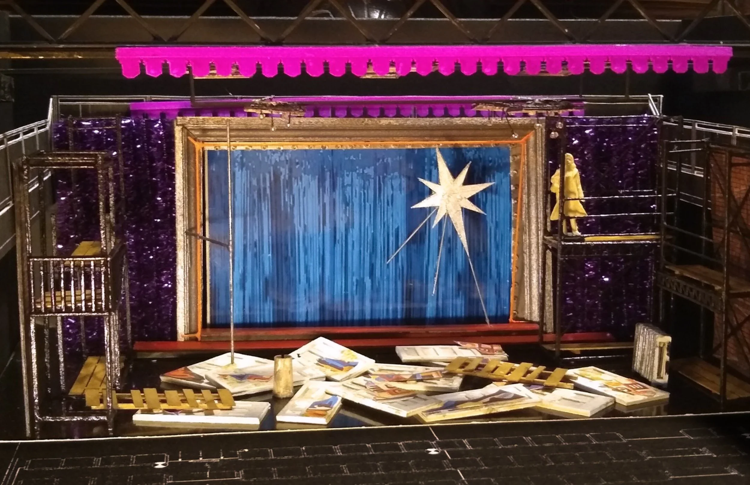 A giant star sits in front of a blue curtain in a gold frame in a sparkling purple wall. A figure stands on a scaffold looking down on detritus covering the floor.