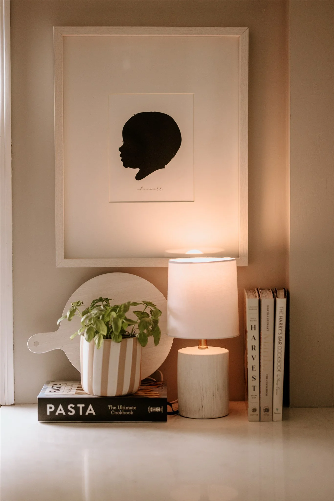 A framed silhouette art piece of a child's profile on the wall. On a surface below, a table lamp, a potted plant with green leaves, a cutting board, and cookbooks titled "Pasta," "Harves," "The Harry's Bar Cookbook," are visible.