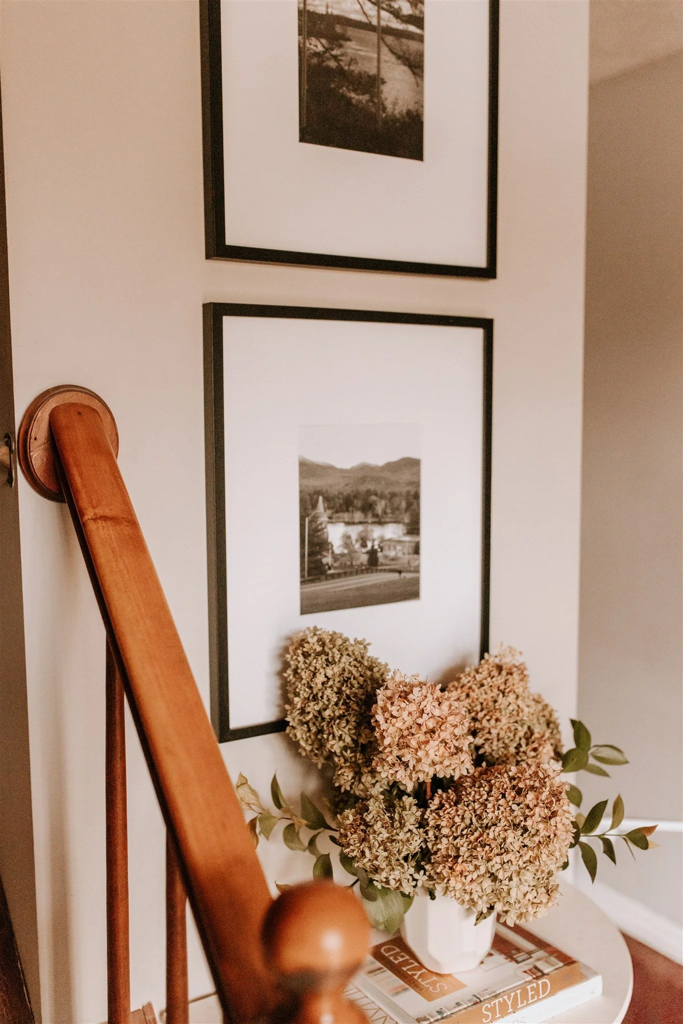 A cozy corner with framed black and white landscape photographs on the wall, a wooden handrail, a vase of dried hydrangeas on a small table, and a stack of magazines.