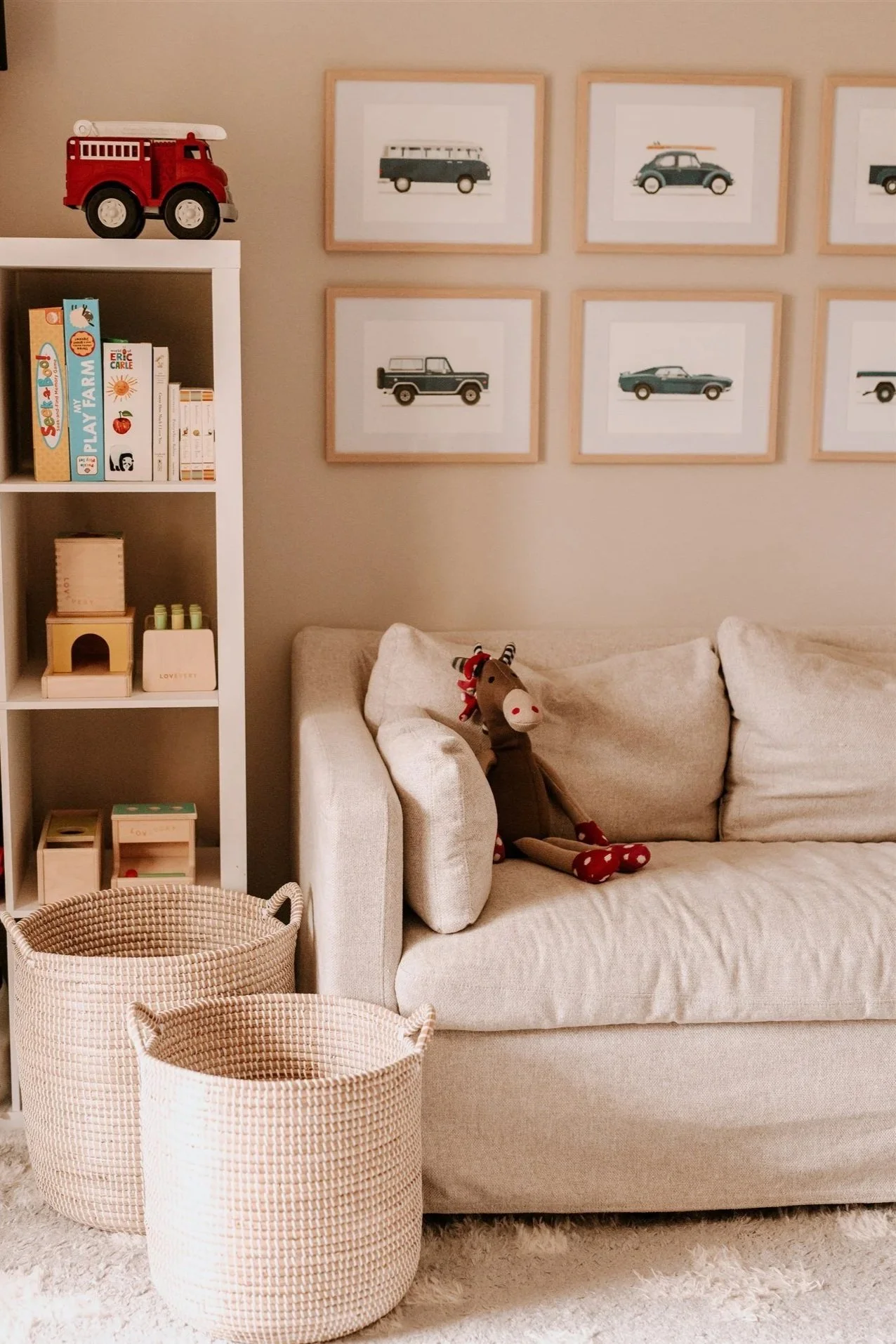 A cozy living room corner with a beige sofa, a stuffed reindeer toy, a white bookshelf filled with children's books and toys, two woven baskets on a plush white rug, and framed artwork of vintage cars and vans on the wall.