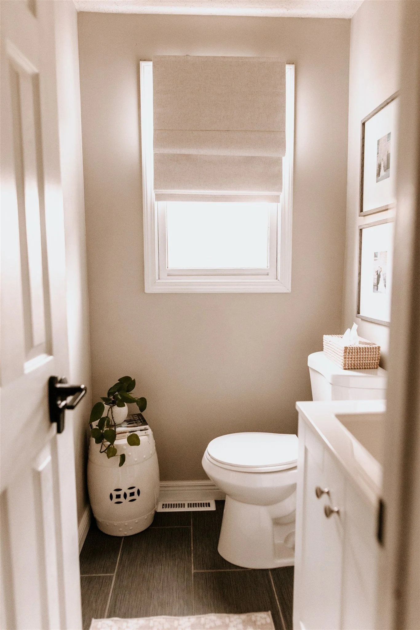 A small, minimalist bathroom with a white toilet, a window with a beige Roman shade, a white cabinet, and a decorative ceramic stool with a plant on top, dark tiled floor, and framed pictures on the wall.