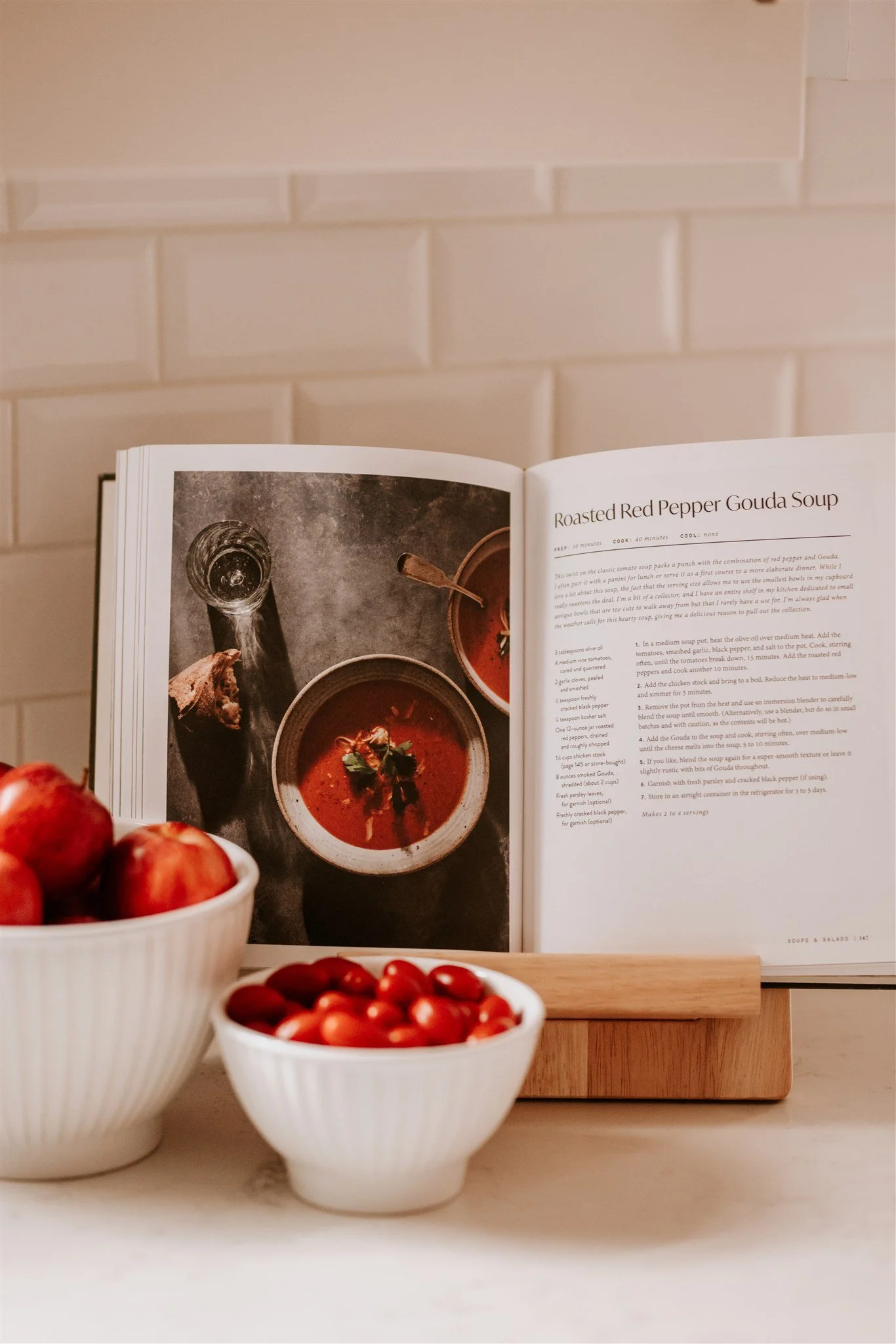 Open cookbook displaying a recipe for roasted red pepper gouda soup, with bowls of tomatoes and small peeled tomatoes in front, set against a white tiled wall background.