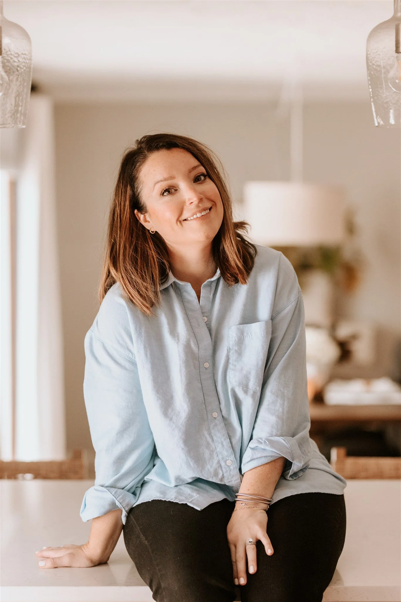A woman with shoulder-length brown hair, smiling, wearing a light blue button-up shirt and black pants, sitting on a kitchen counter.
