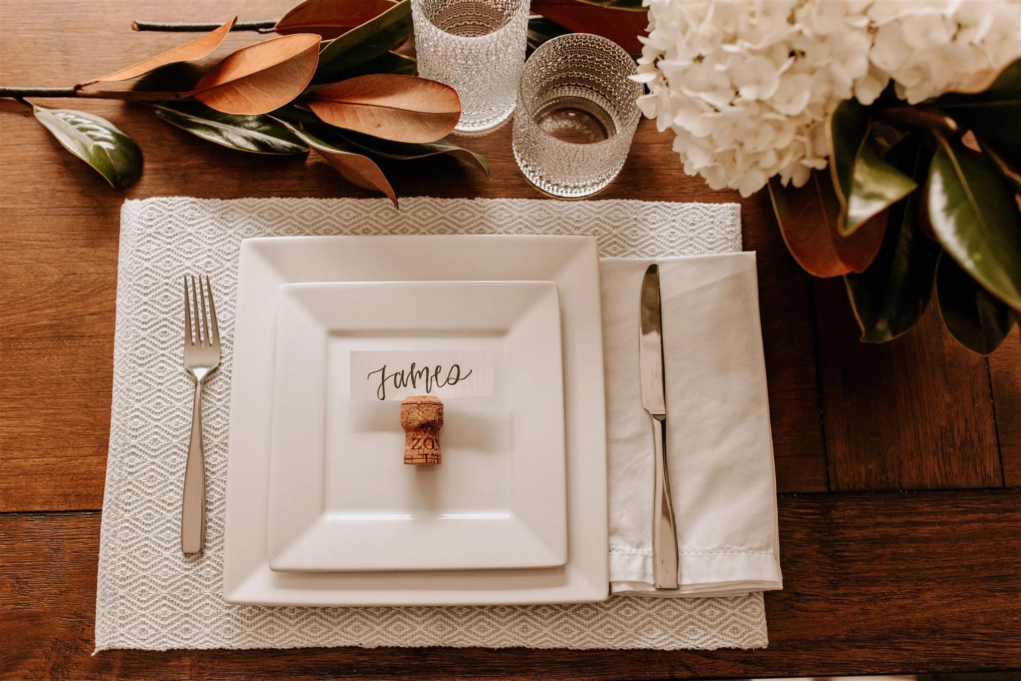 Elegant dining table set with white square plates, a place card with the name "James" and a cork, a fork on the left, a knife on the right, a white napkin underneath, two textured glasses, and a floral centerpiece with white flowers and green and bro