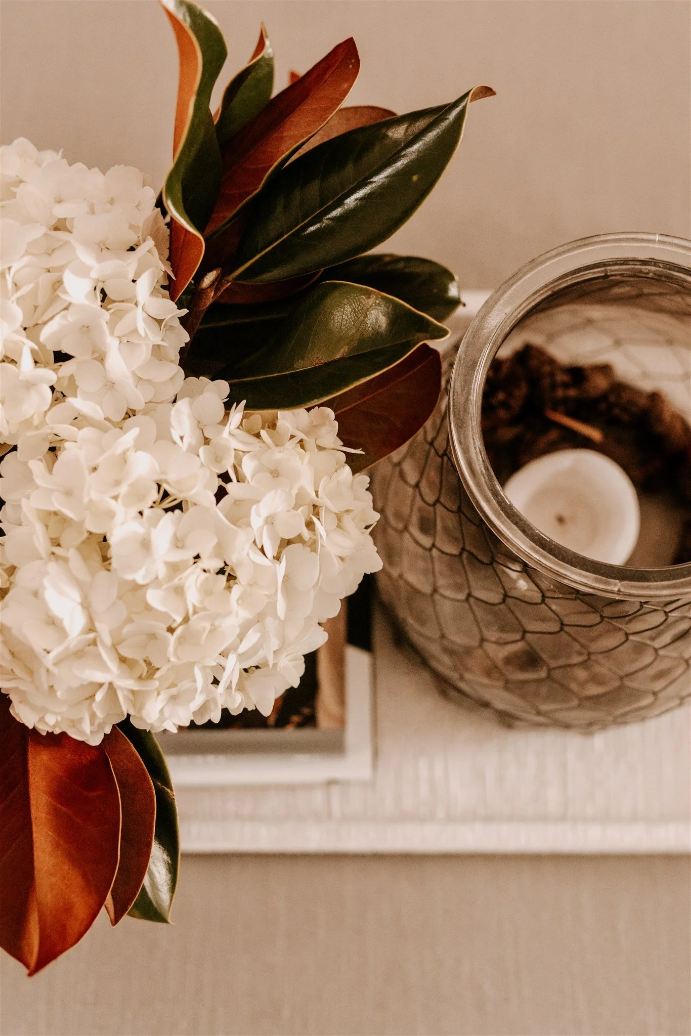 Close-up of a white hydrangea flower bouquet with dark green and brown-tinted leaves next to a glass candle holder with a white candle and some decorative brown objects inside.
