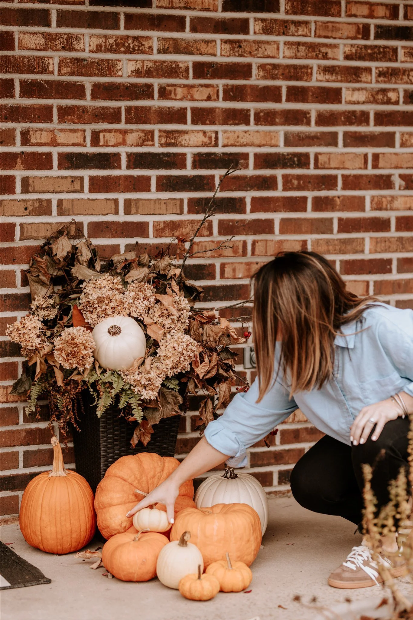 A woman is arranging pumpkins and gourds of various sizes and colors near a large floral arrangement featuring dried leaves, hydrangeas, and a white pumpkin, against a red brick wall.