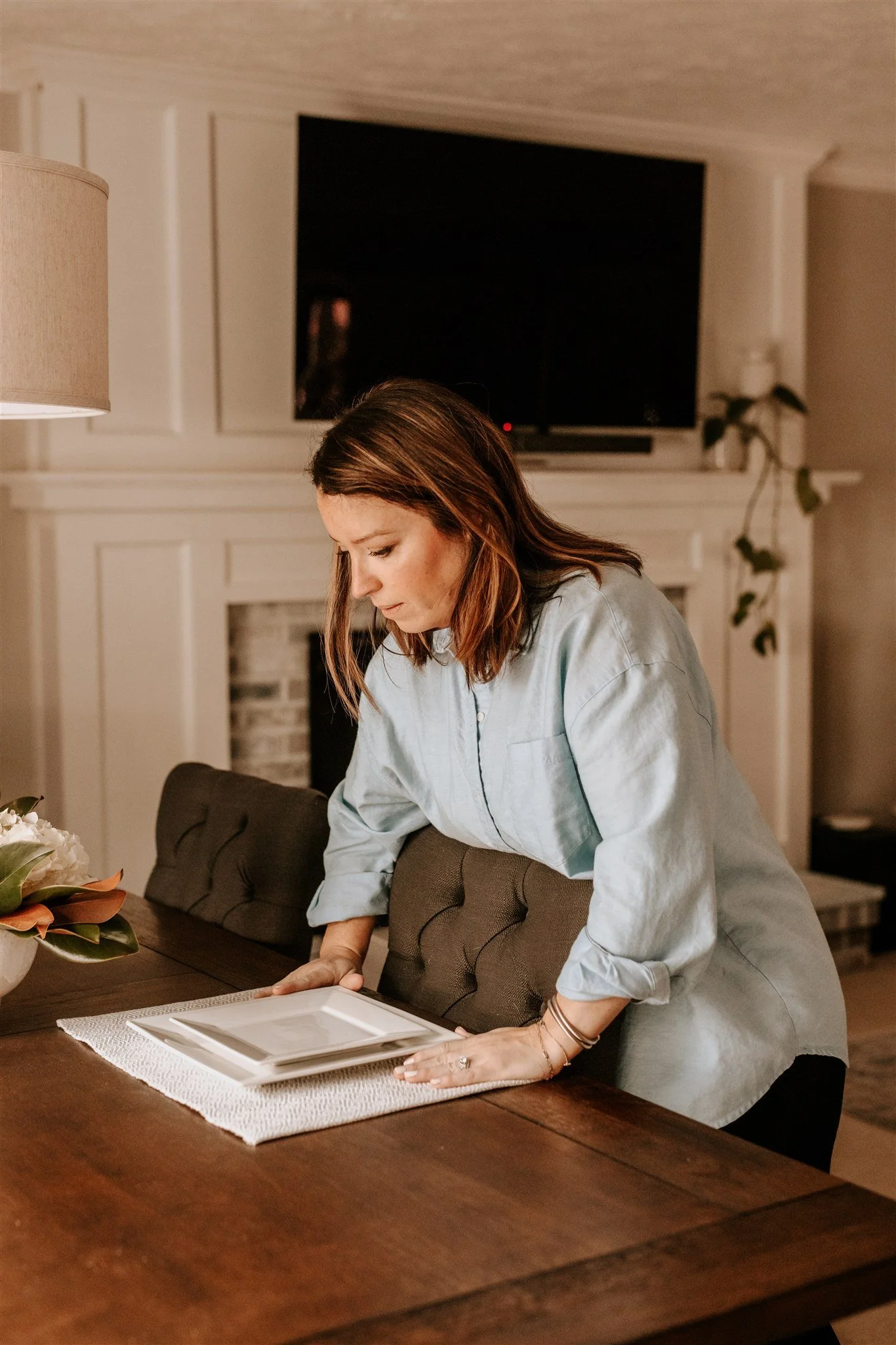 A woman with shoulder-length brown hair wearing a light blue shirt is leaning over a dining table with a white placemat and a flower arrangement on it, arranging or picking up a white rectangular tray or plate.