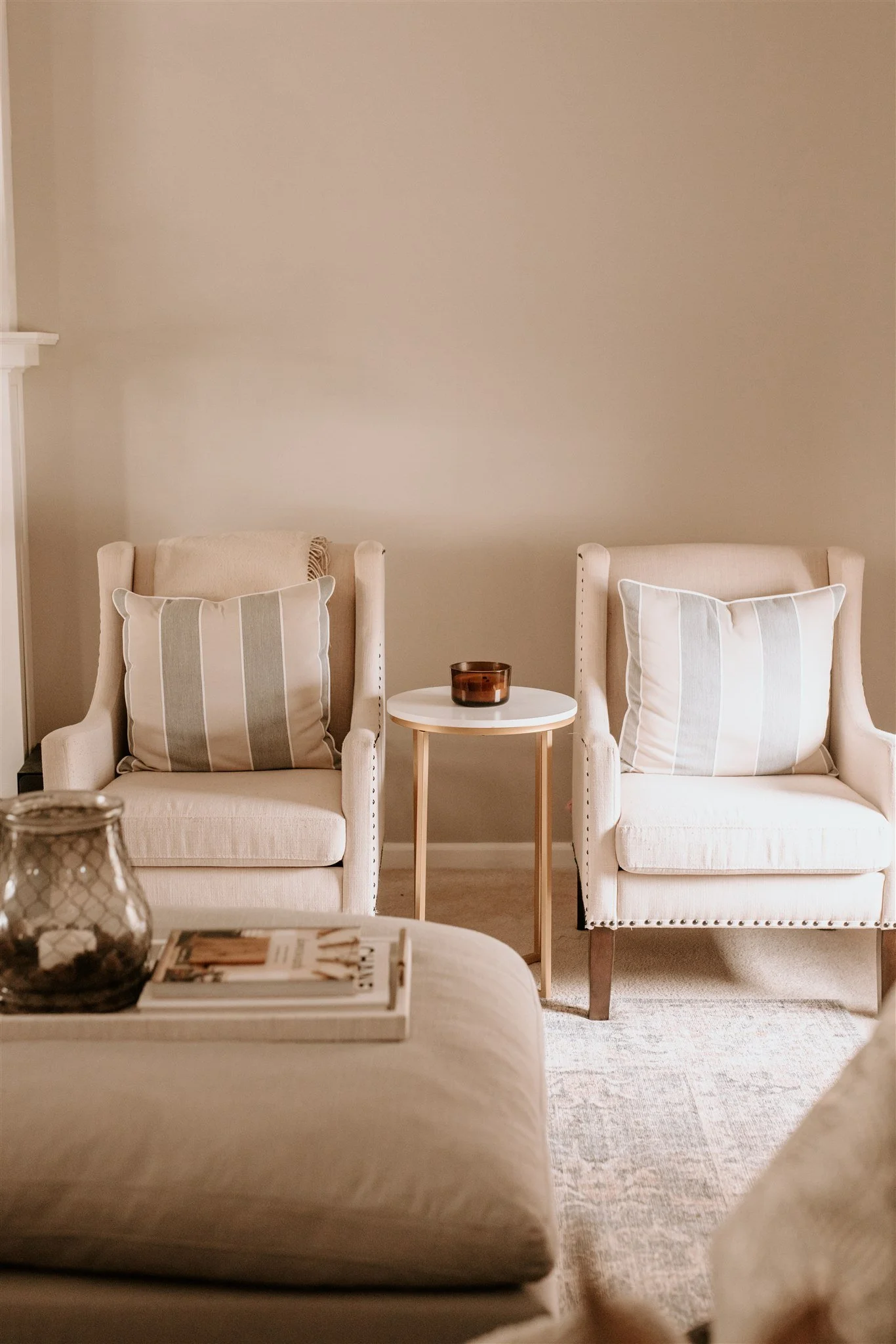 Two beige armchairs with striped pillows, separated by a small round white side table with a brown glass container on top. Part of a beige ottoman with a magazine and a candle holder is visible in the foreground. The wall behind is plain and light-co