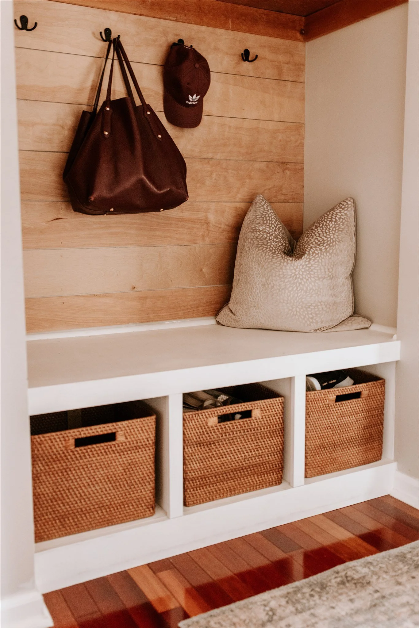 Wall with hooks holding a brown handbag and cap, a cushioned pillow on a white shelf with built-in cubbies below, containing woven baskets with shoes, next to a wooden wall and floor.