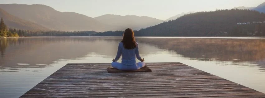 Woman practicing meditation on a dock overlooking a peaceful mountain lake, representing mindfulness, relaxation, and emotional well-being.