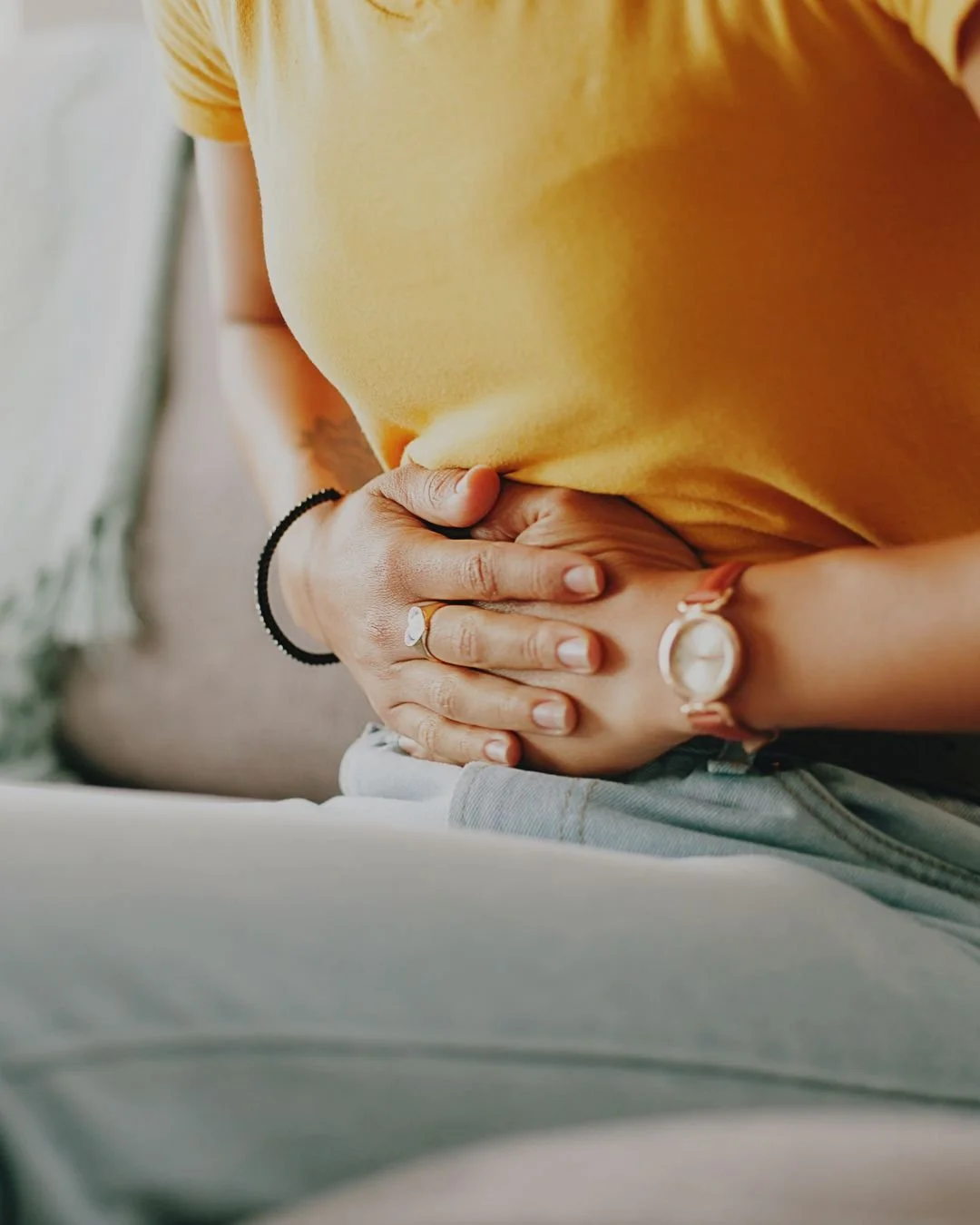 A person clutching their stomach, possibly indicating pain or discomfort, wearing a yellow shirt and accessories including a ring, watch, and black hair tie.