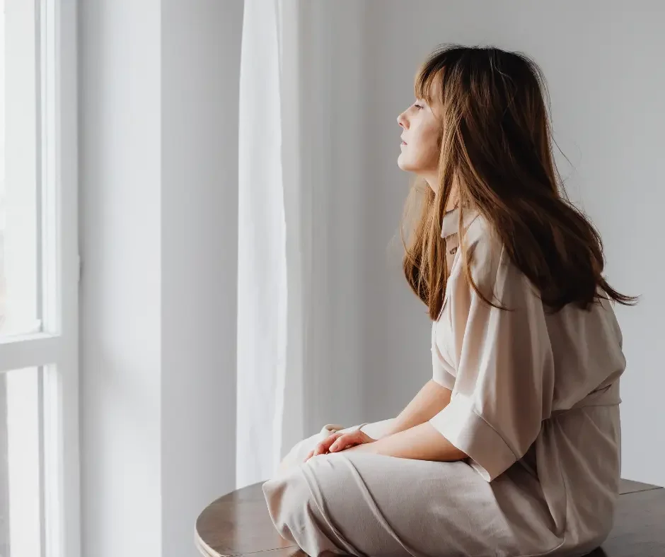 Woman sitting beside a window with a thoughtful expression, representing the emotional and physical impact of living with chronic pelvic pain and endometriosis.