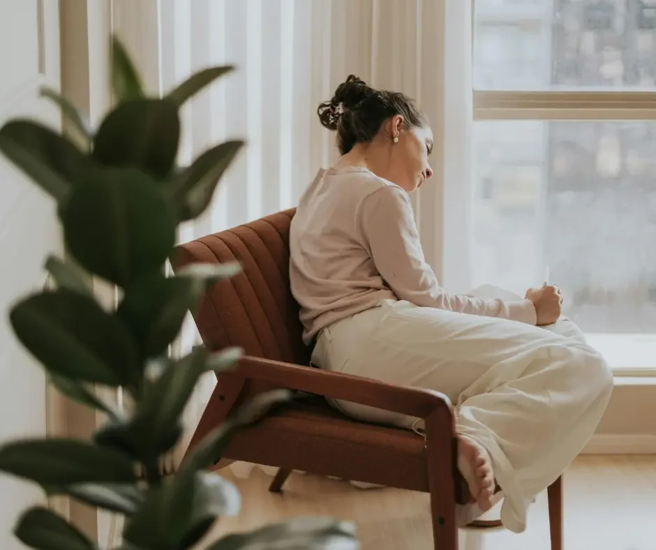 Woman sitting quietly in a chair by a window, appearing reflective and emotionally overwhelmed, representing the emotional and mental symptoms associated with PMDD.
