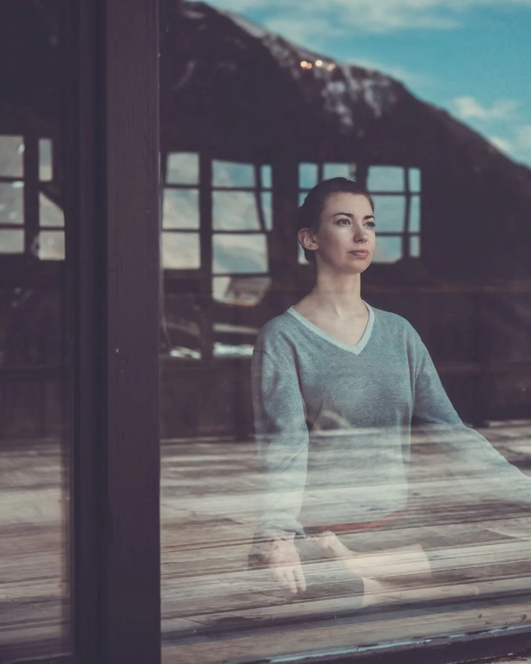 A woman with dark hair sitting indoors, gazing out a window with a wooden frame. There are mountains and a blue sky with clouds visible outside.